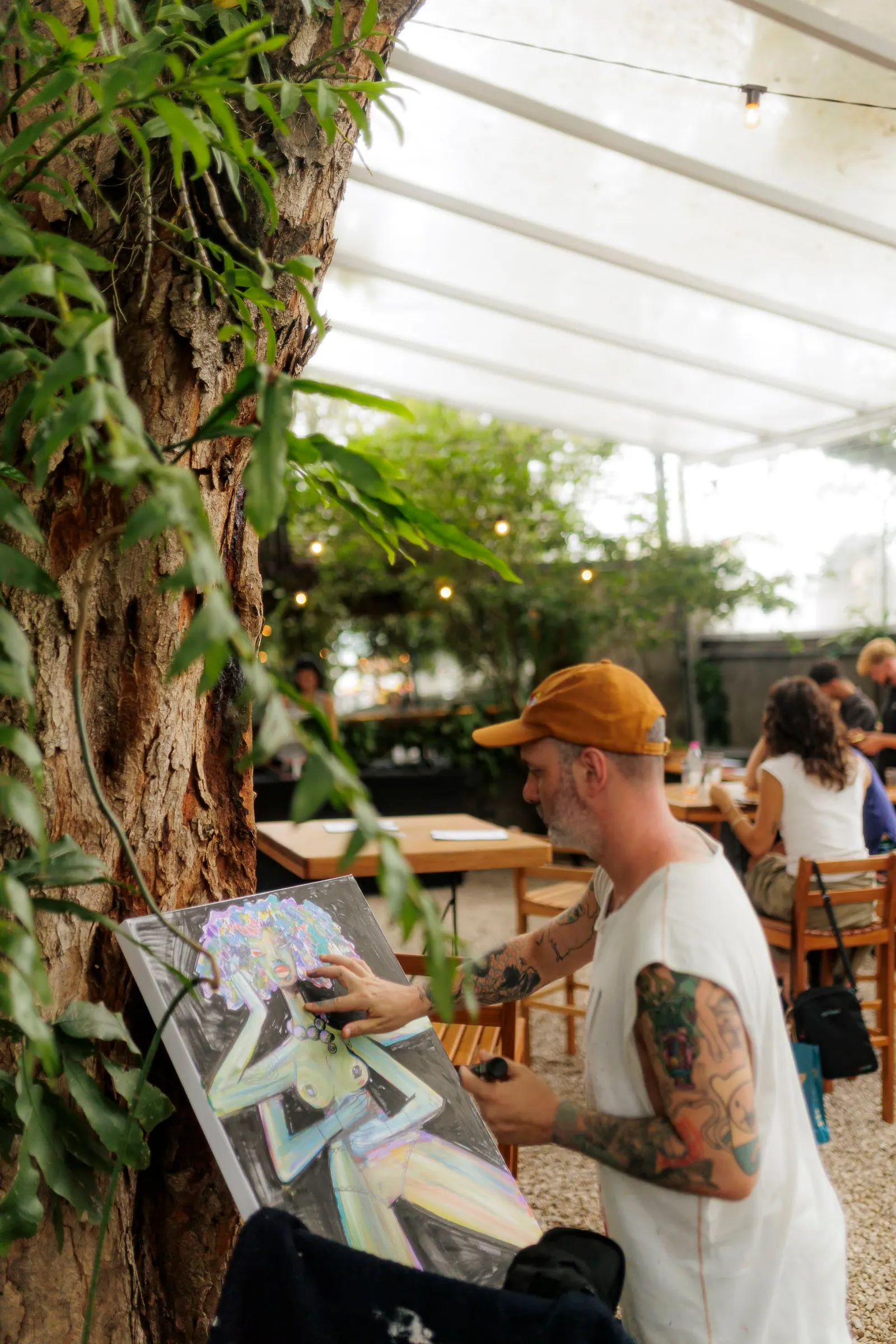 Artista de braços tatuados pinta um quadro colorido apoiado numa árvore, terraço coberto de um evento de garimpo