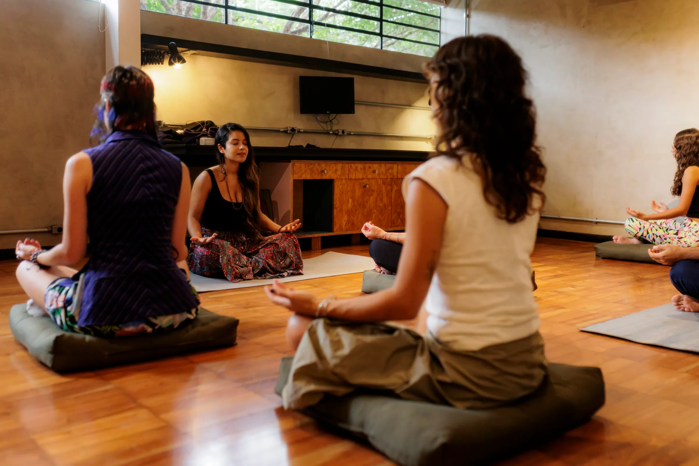 Roda de meditação sentada em almofadas no chão, mãos em posição de oração e olhos fechados