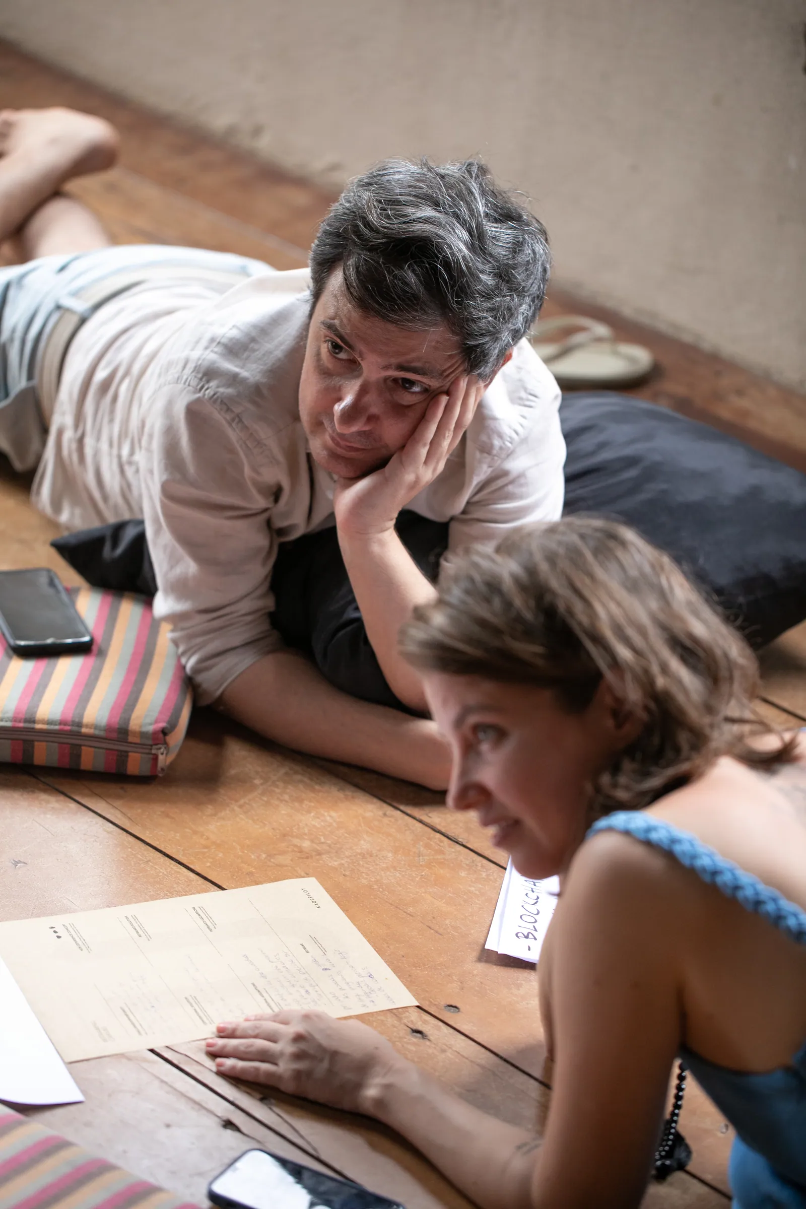 Dois amigos deitados no chão de madeira conversam sobre papéis e o celular, clima descontraído