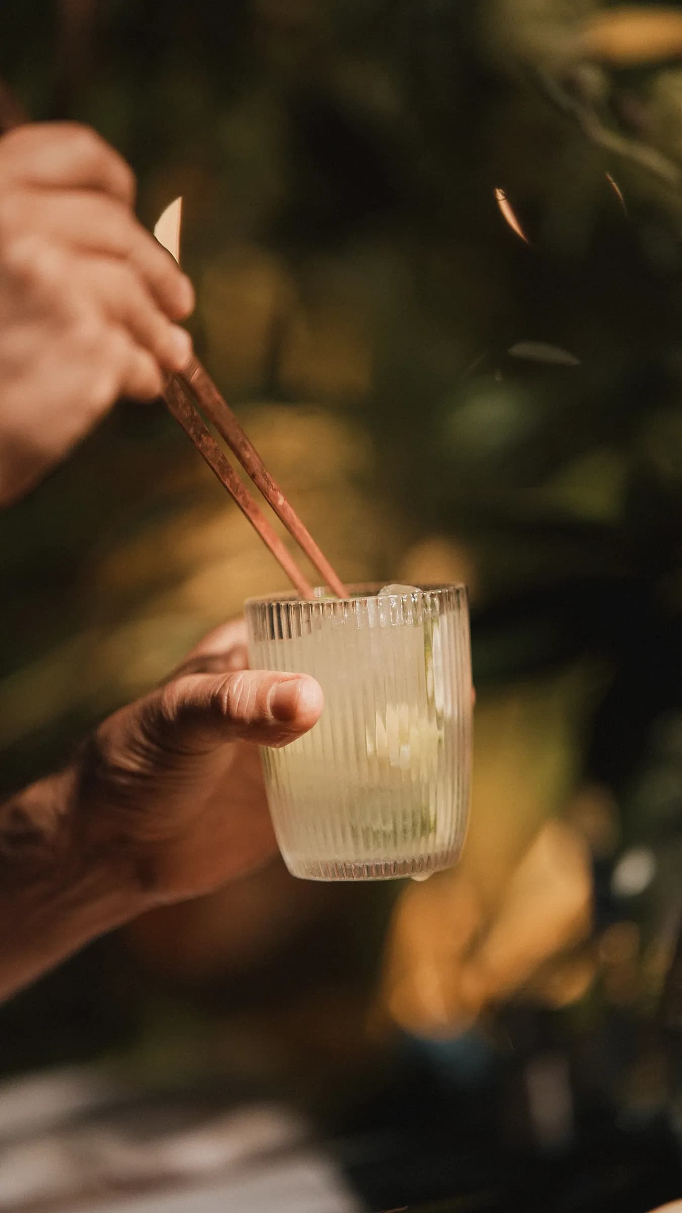Mão mexendo um drink claro com colher longa, entre plantas, luz noturna