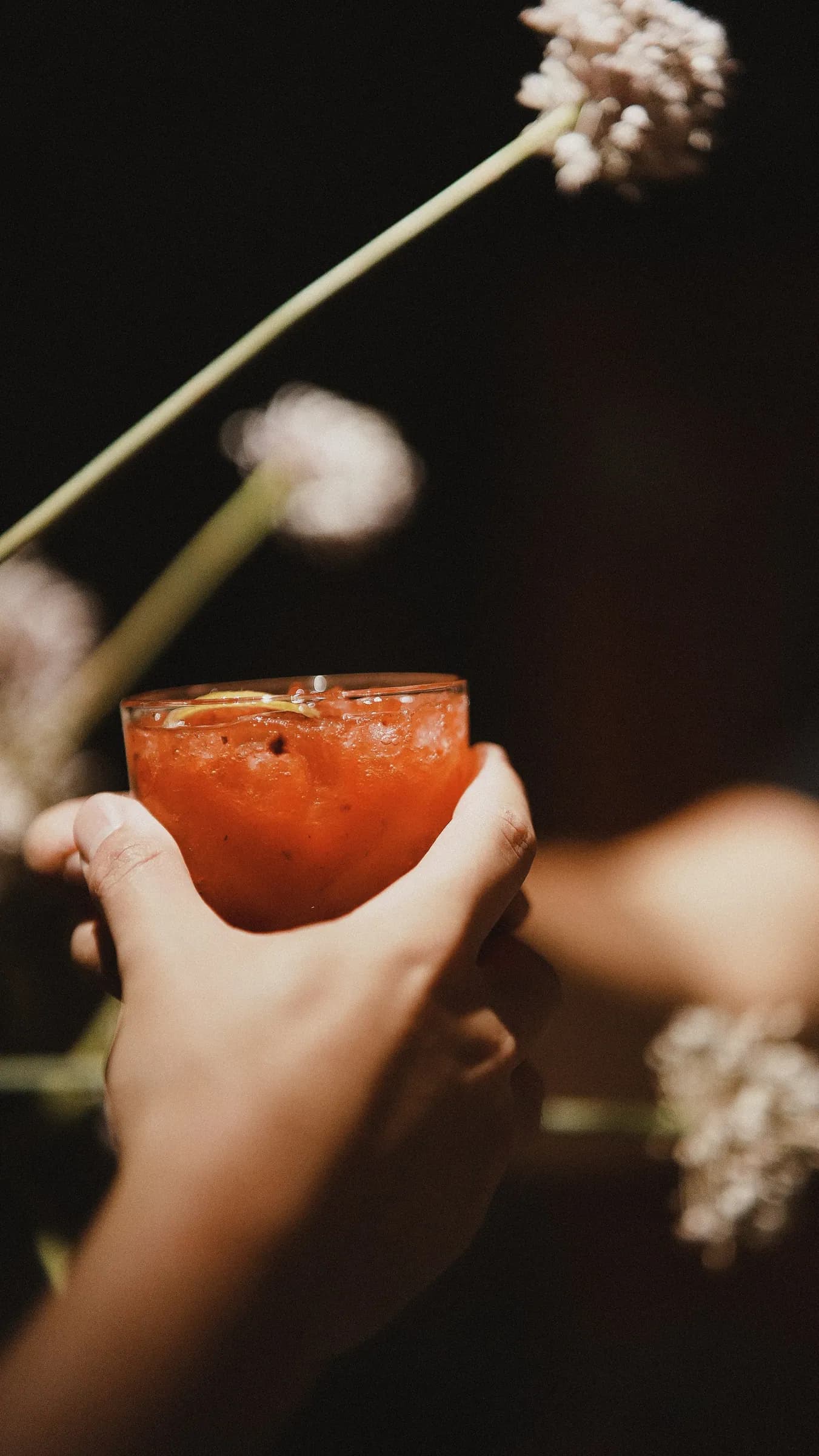 Mãos passando um copo de drink vermelho entre flores brancas