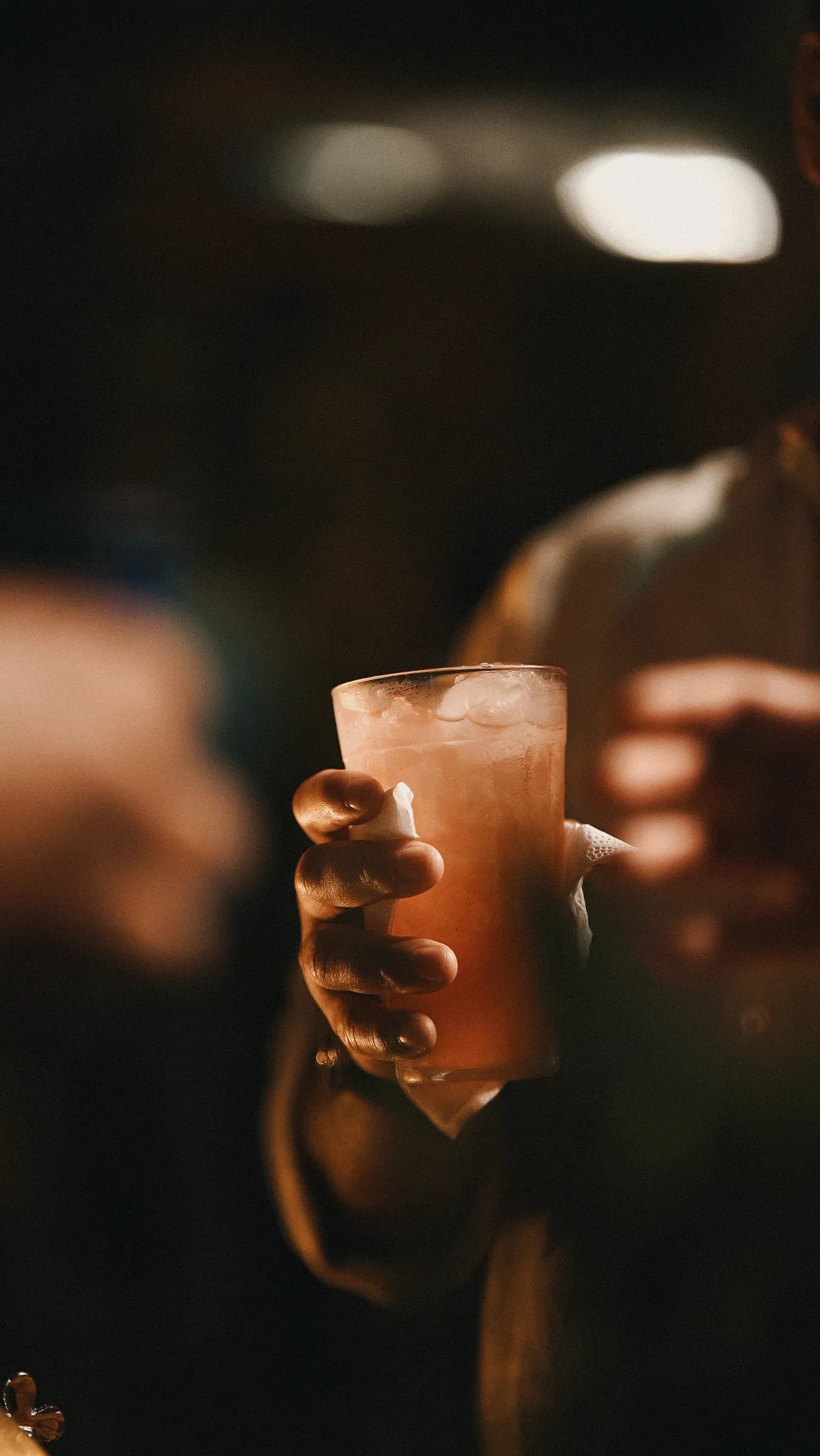 Convidado segurando copo de drink rosado, luzes da festa desfocadas ao fundo