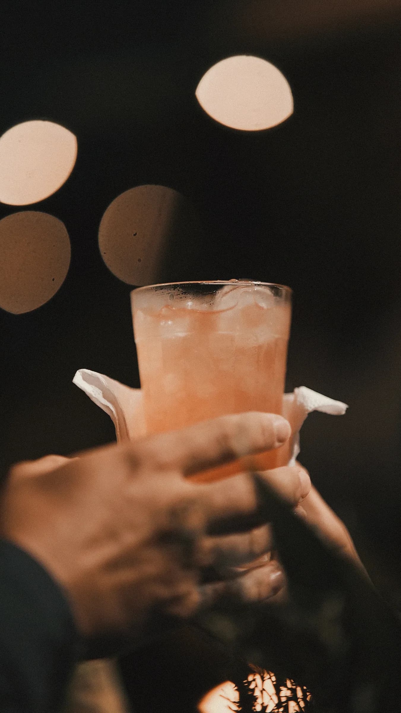 Mãos brindando com copo de drink rosado gelado, luzes da festa ao fundo