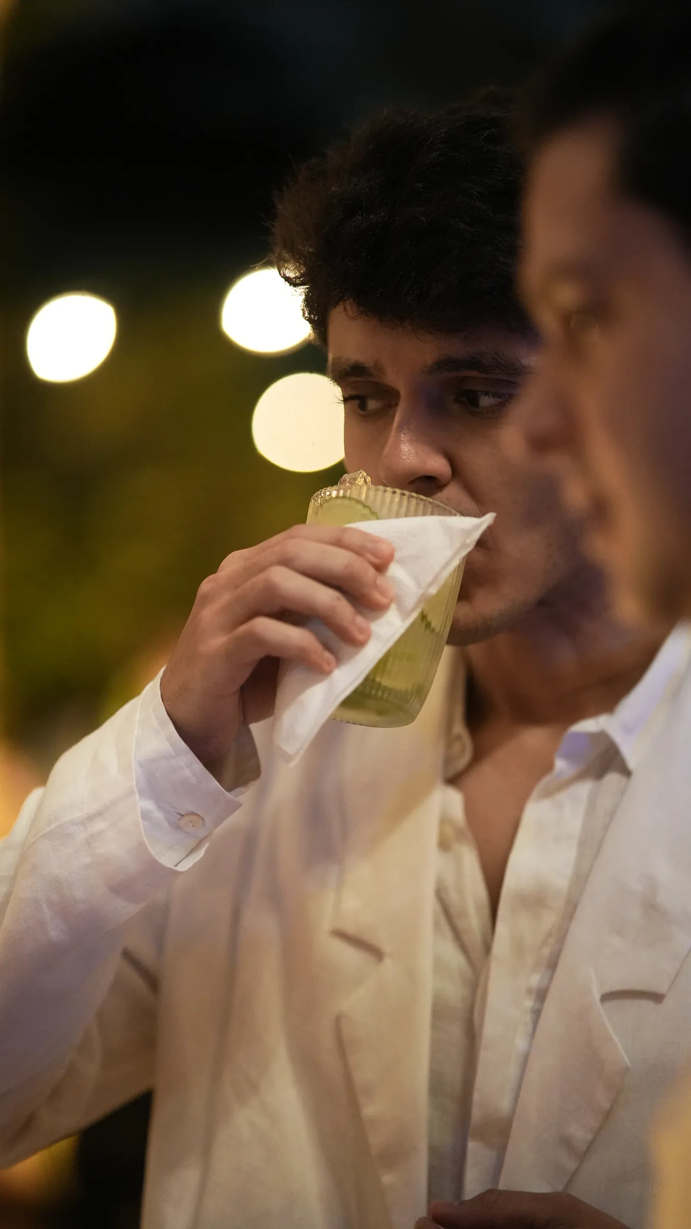 Dois convidados brindando e bebendo juntos, sorrindo, luzes da festa ao fundo