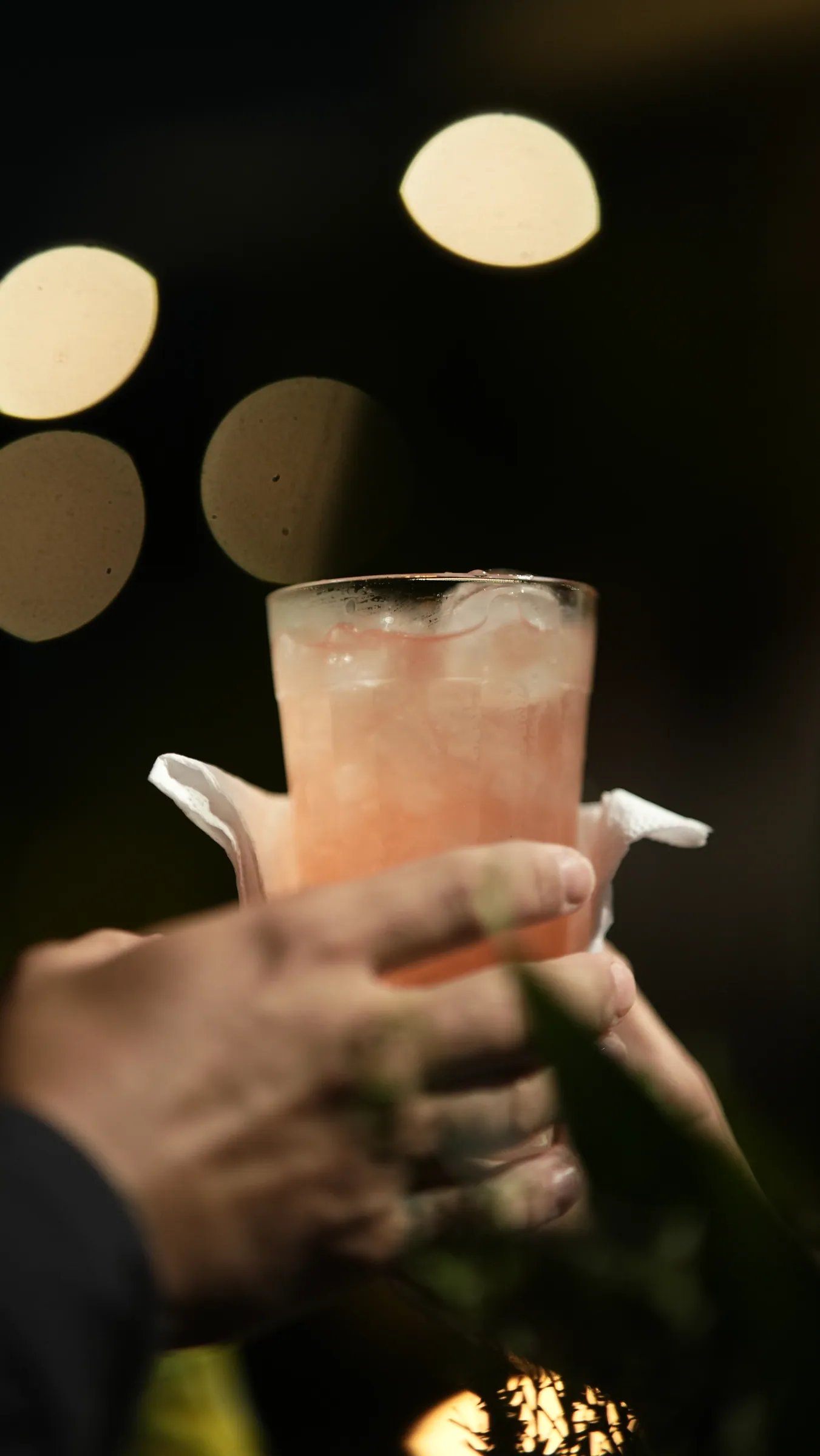 Mãos brindando com copo de drink rosado, luzes da festa desfocadas ao fundo