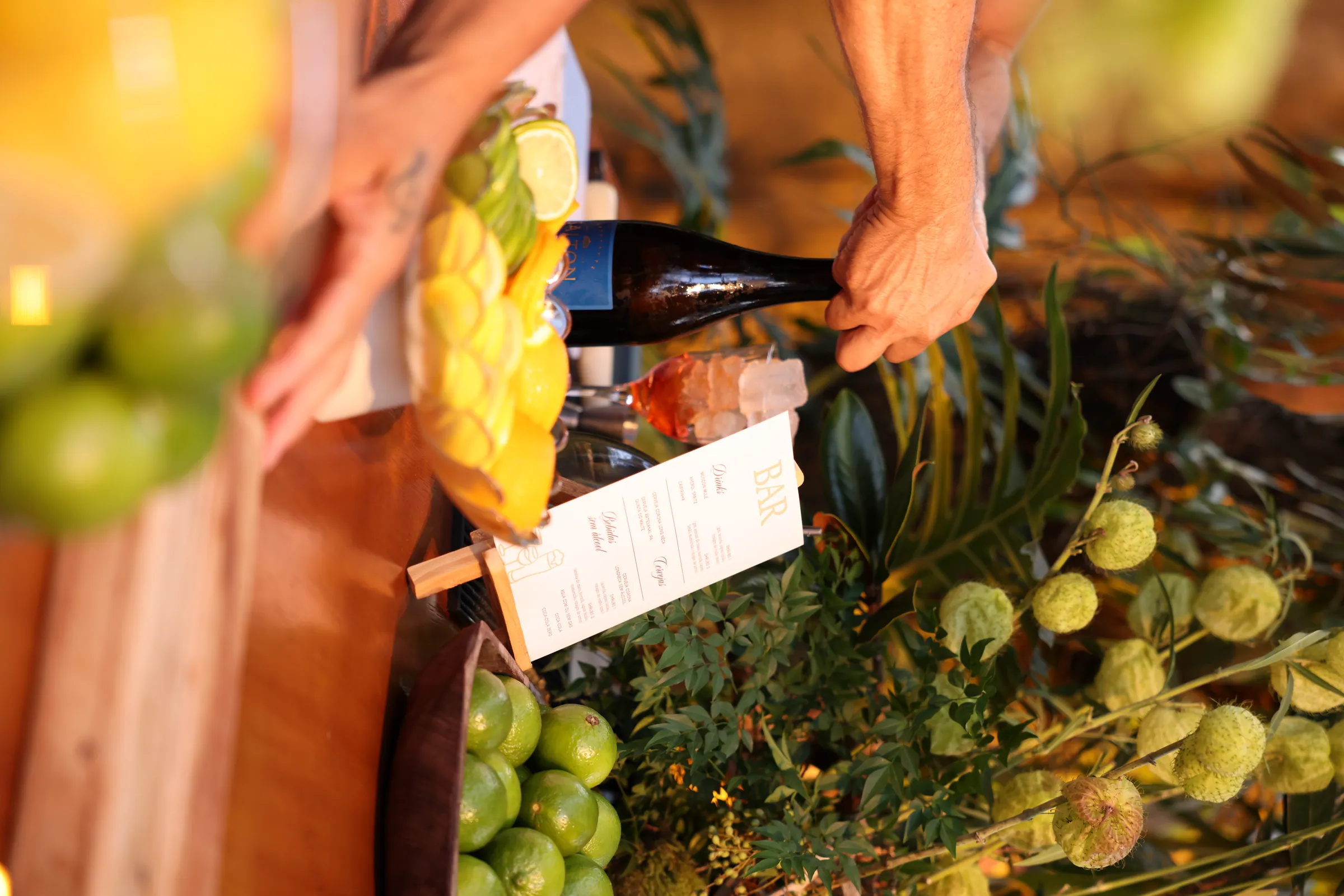 Mão pegando garrafa em meio à decoração de frutas e folhagens tropicais