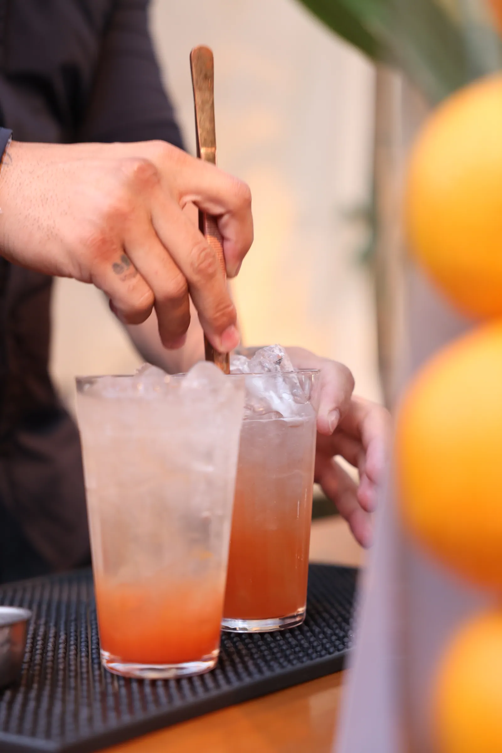 Bartender mexendo drink com colher longa, dois copos com gelo, luz alaranjada ao fundo