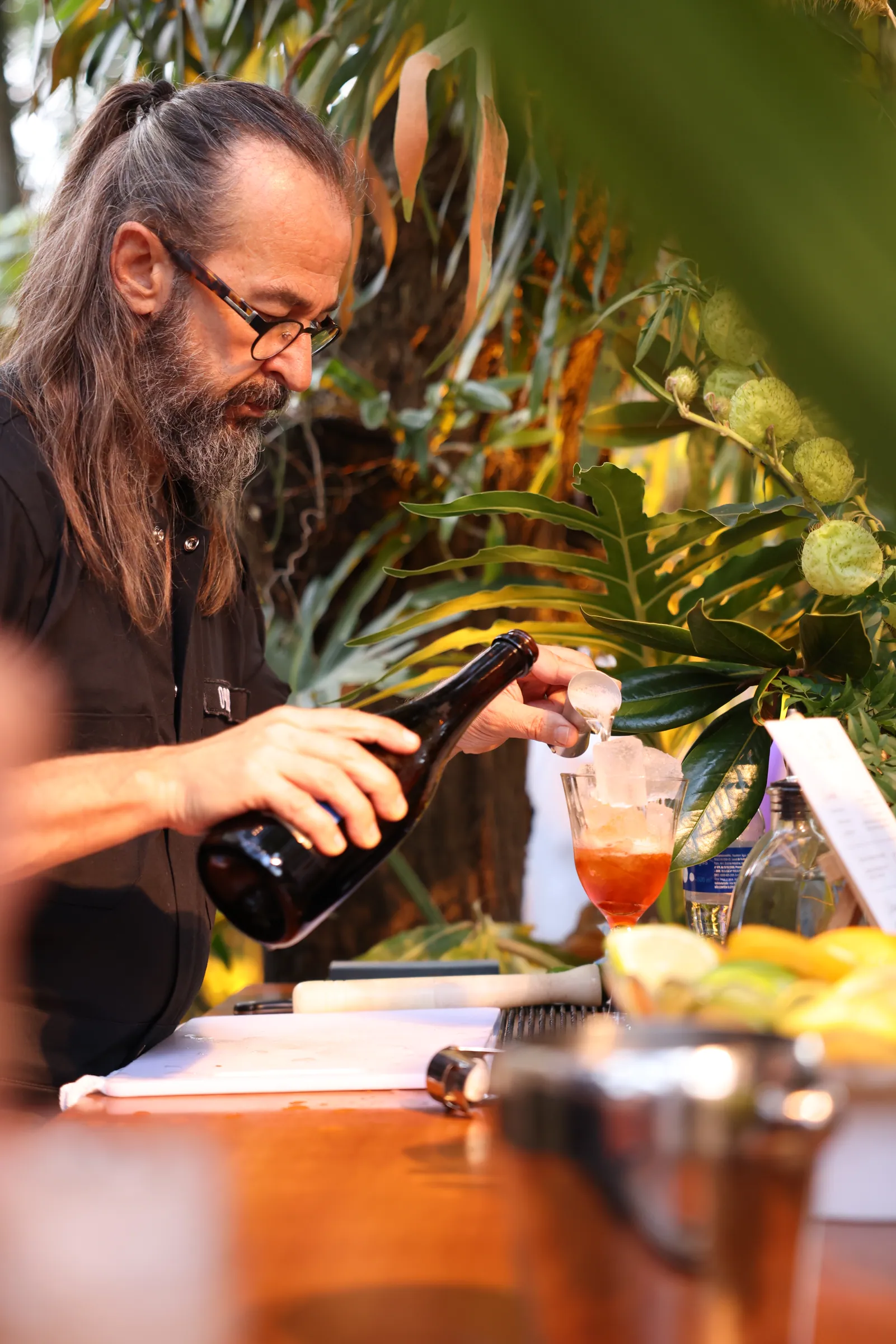 Bartender de barba e cabelo longo preparando drinks na estação, cercado de folhagem tropical