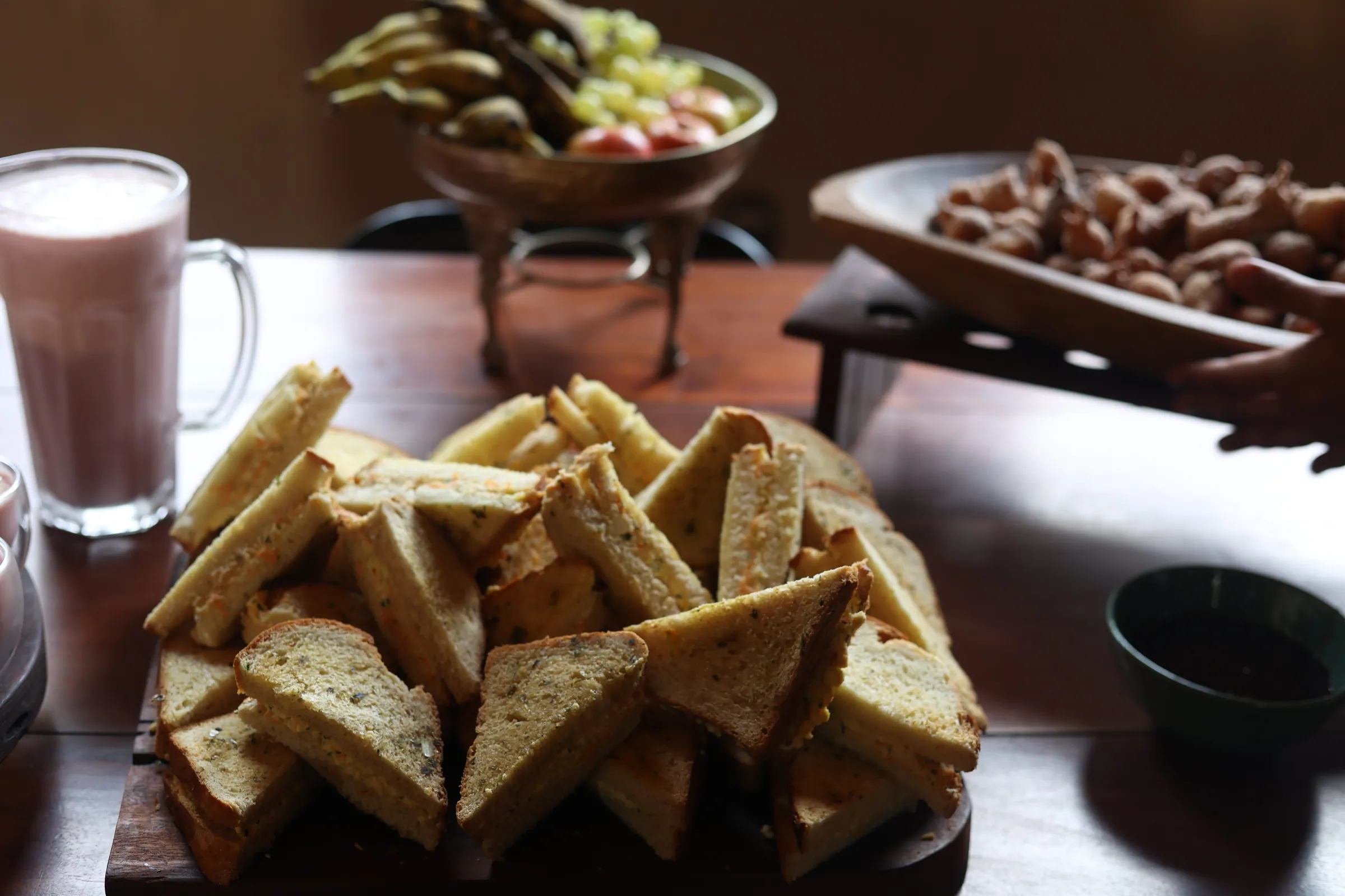 Mesa de coffee break com sanduíches naturais cortados em triângulo, tigela de frutas ao fundo e copo de vitamina rosada
