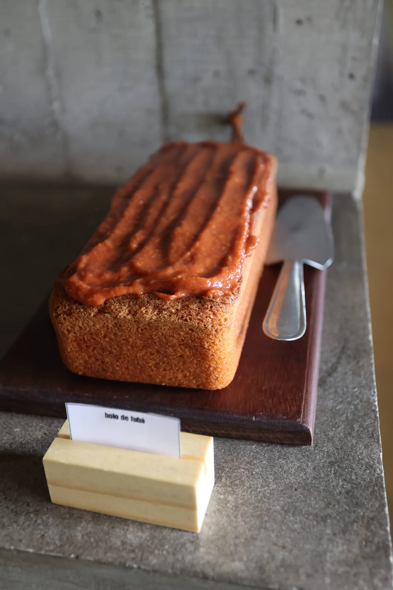 Bolo de fubá inteiro com cobertura de doce de leite listrada, faca de bolo ao lado e plaquinha escrita "bolo de fubá"