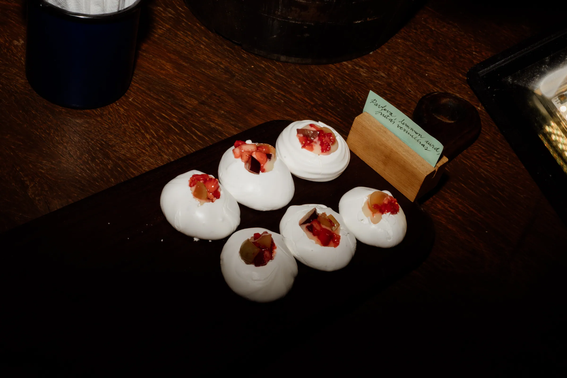 Seis mini pavlovas com frutas vermelhas e uva sobre tábua escura, com cartão escrito à mão descrevendo a sobremesa