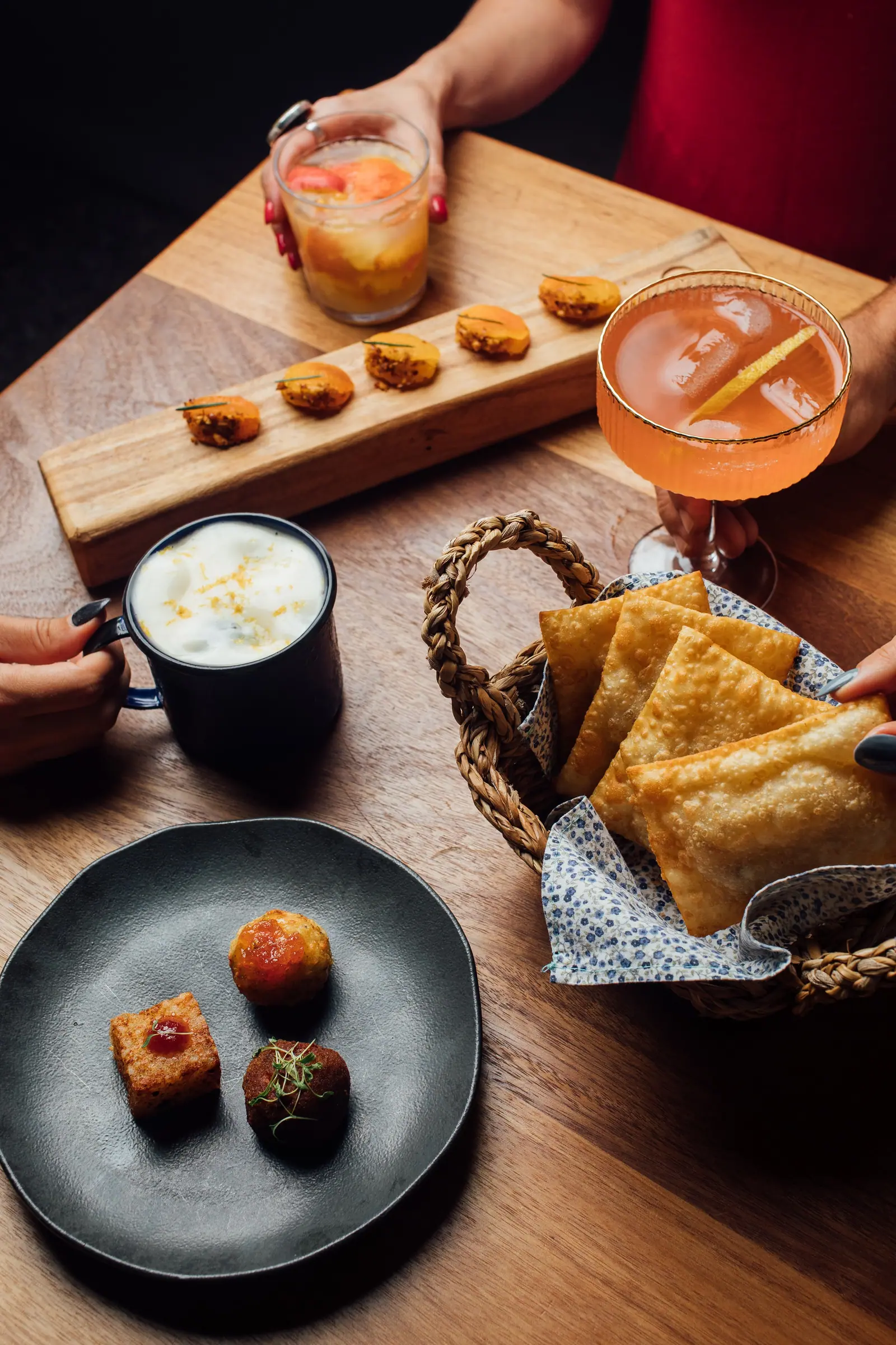 Convidados servindo-se de uma cesta de chips crocantes e provando croquetes e damascos glaceados numa mesa de madeira, com drinks coloridos em mãos