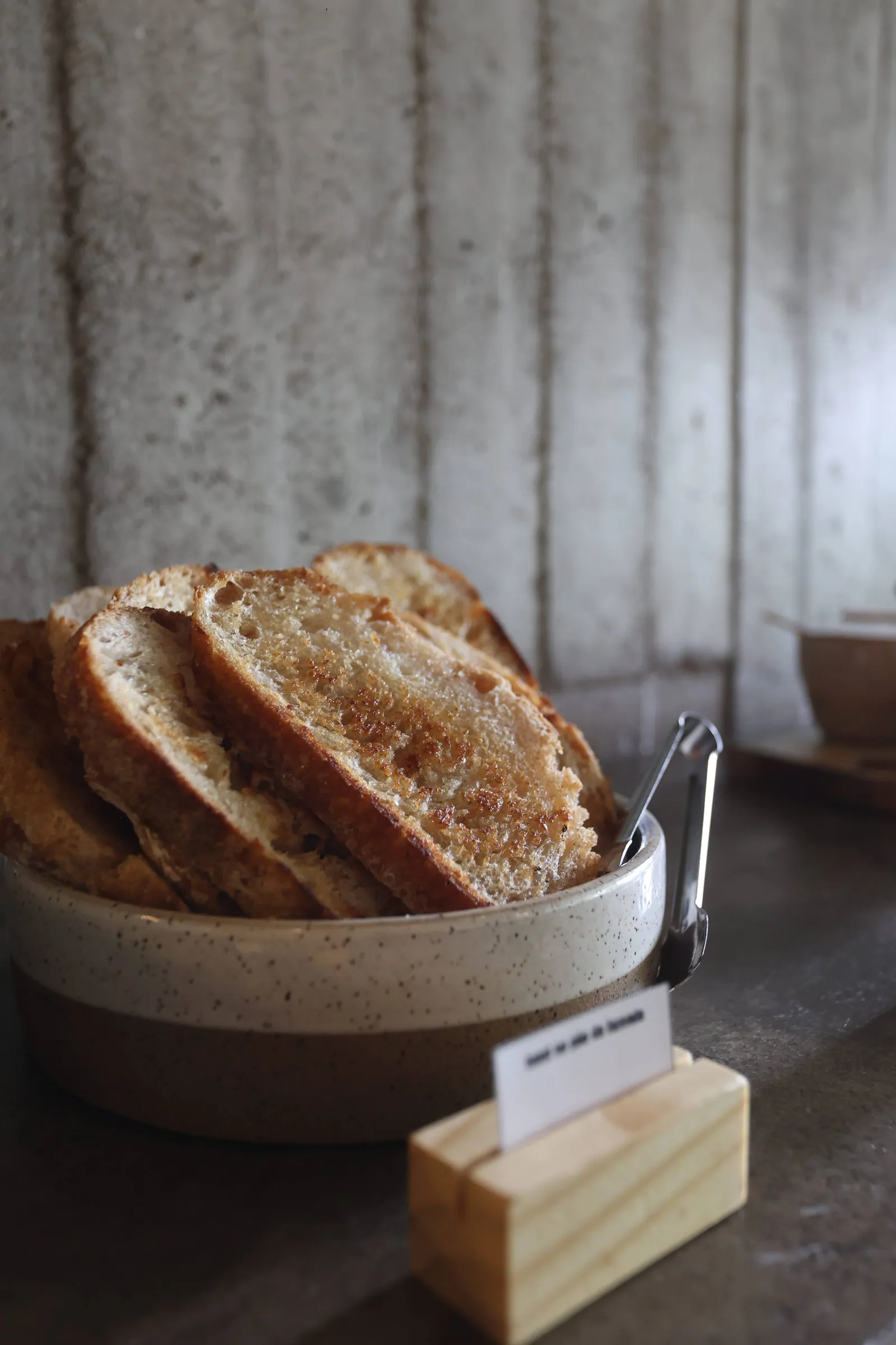 Cesta com fatias de pão tostado e uma plaquinha de identificação, luz suave contra parede de concreto