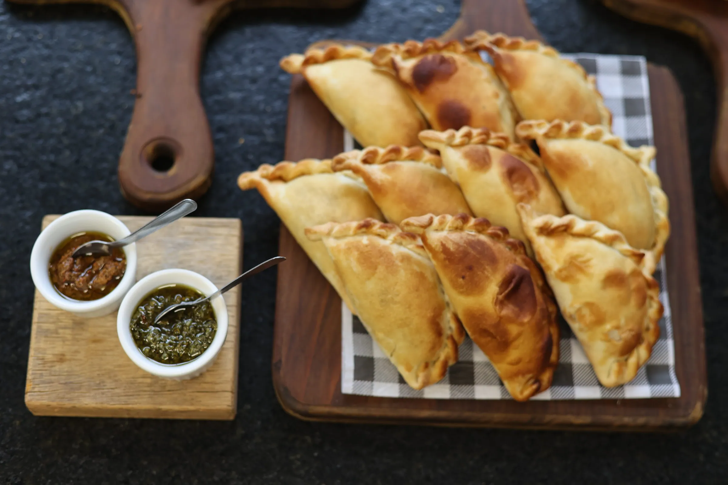 Fileira de empanadas assadas sobre tábua de madeira, acompanhadas de dois molhos em potinhos brancos
