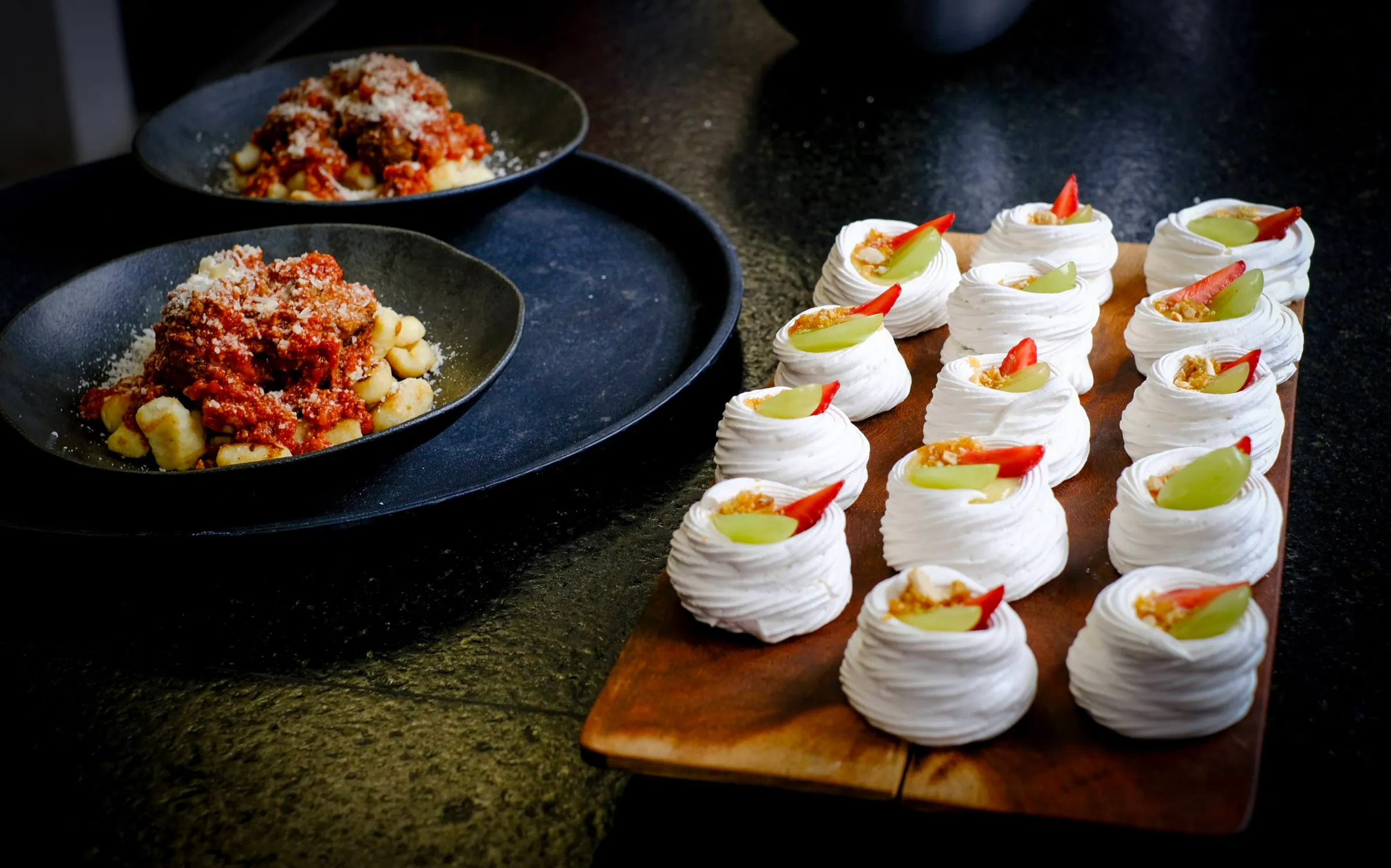 Dois pratos de nhoque ao sugo com parmesão ralado ao lado de uma fileira de mini pavlovas com uva e morango