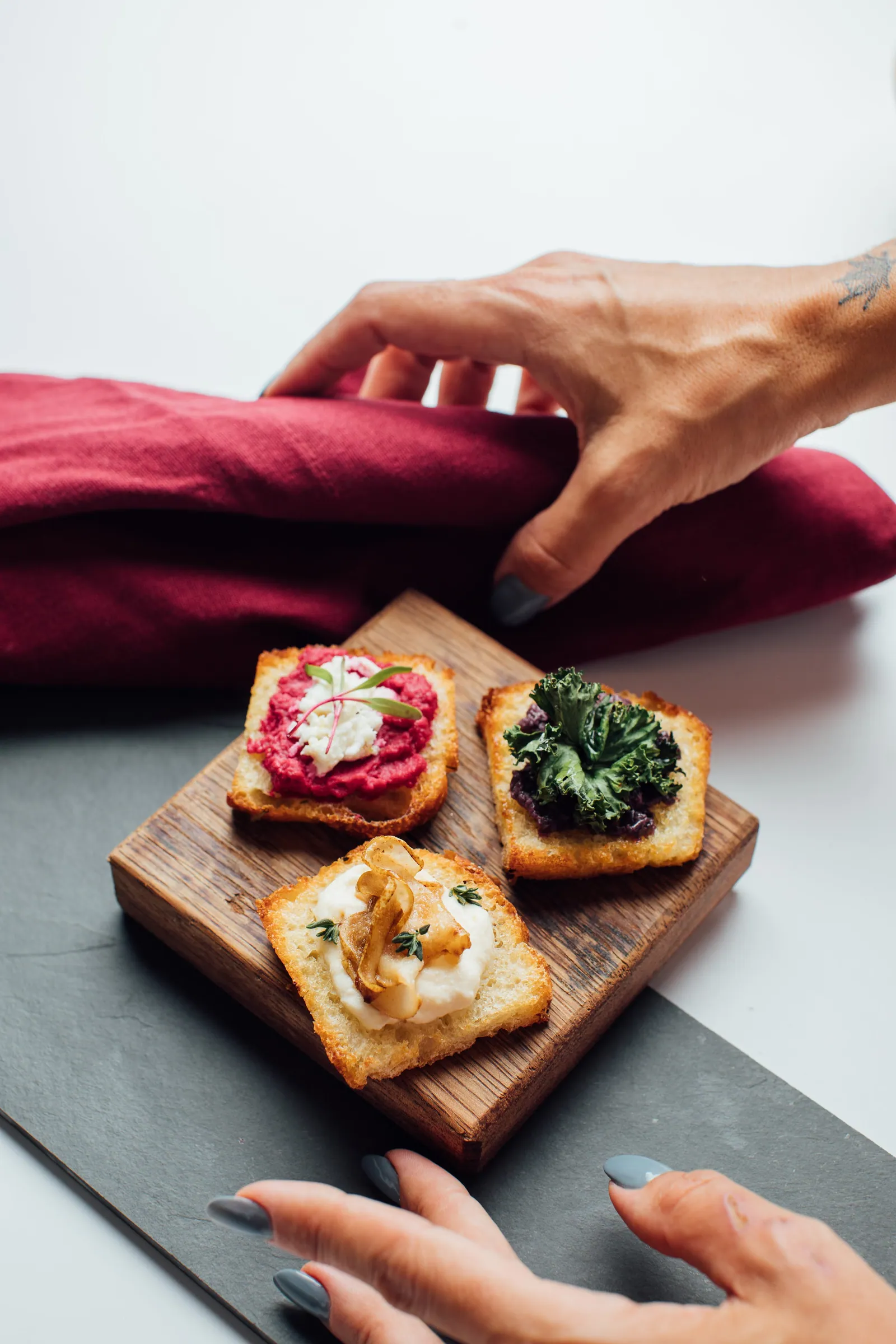 Torradas com três coberturas diferentes numa tábua de madeira — patê de beterraba com queijo cremoso, couve roxa refogada e cogumelo salteado com creme — sendo servidas à mesa