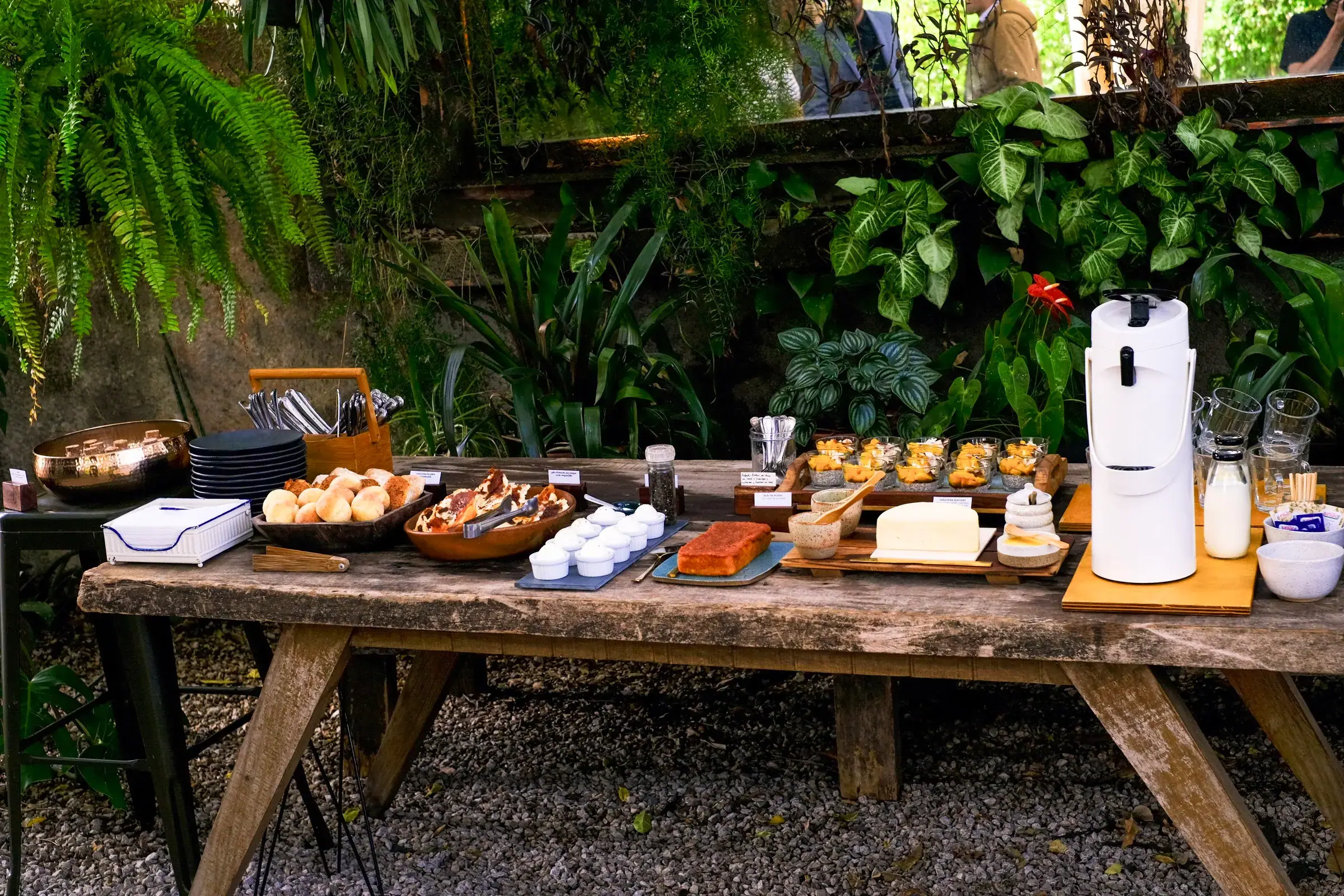 Mesa de buffet montada em jardim, com pães, queijos, geleias e jarra de leite sobre madeira rústica, cercada de folhagem