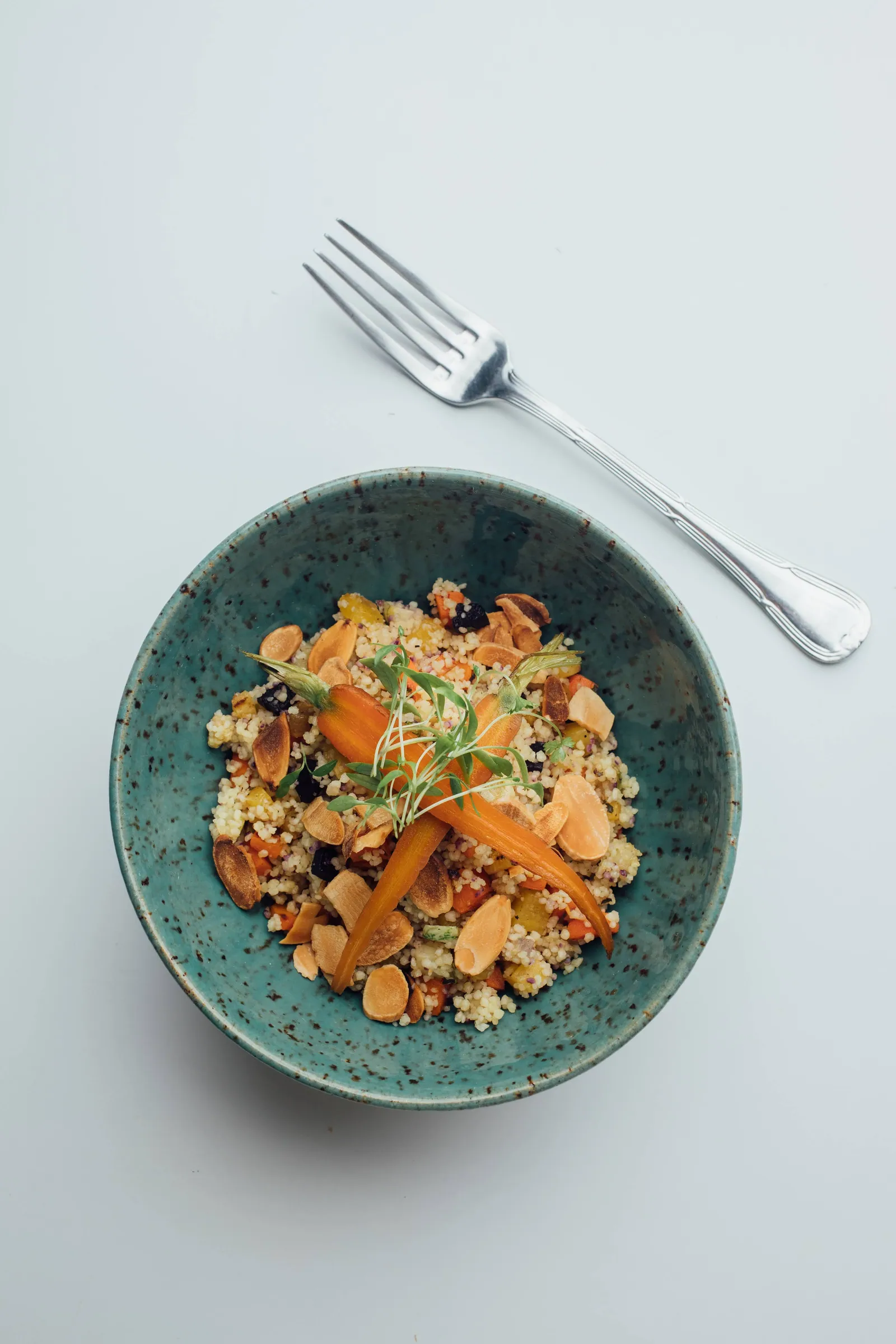 Salada de cuscuz com cenouras assadas, frutas secas e lascas de amêndoa, servida numa tigela de cerâmica azul-esverdeada ao lado de um garfo