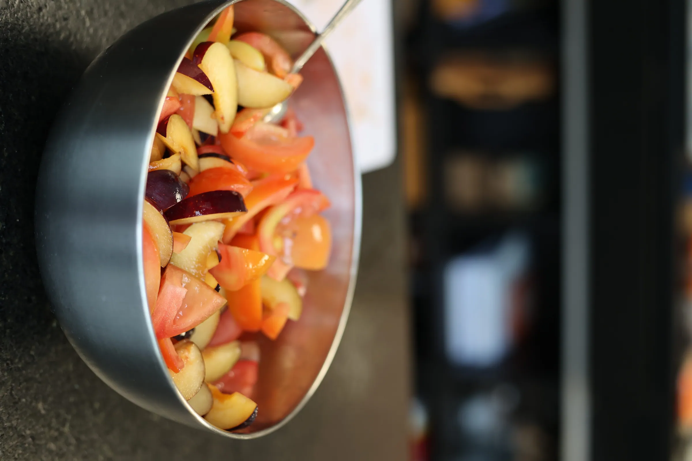 Tigela de salada de frutas com ameixa, tomate e laranja fatiados, colher apoiada na borda