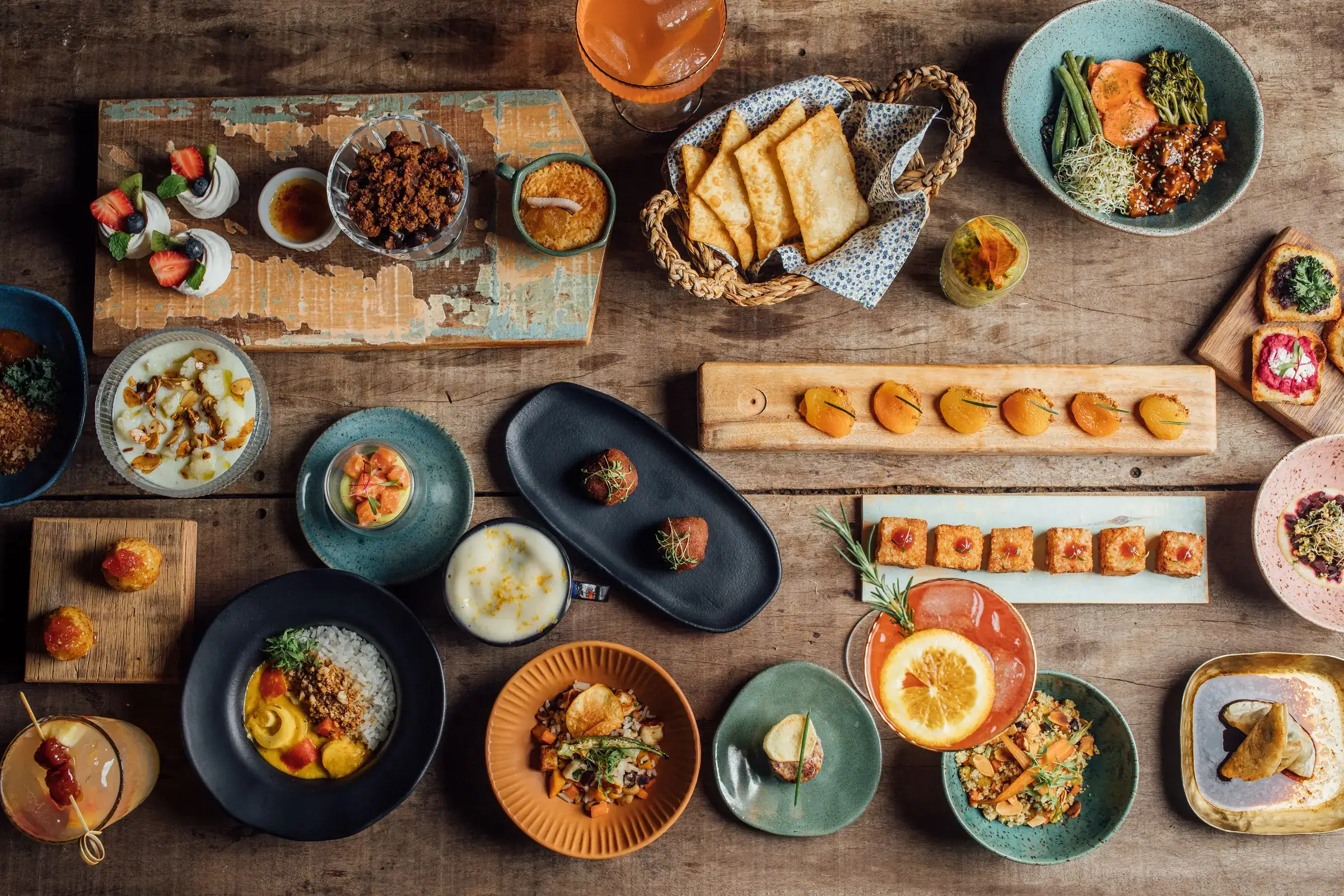 Vista aérea de uma mesa farta com dezenas de pratos variados — curry, croquetes, damascos, canapés, drinks e sobremesas — típica de um evento com cardápio degustação