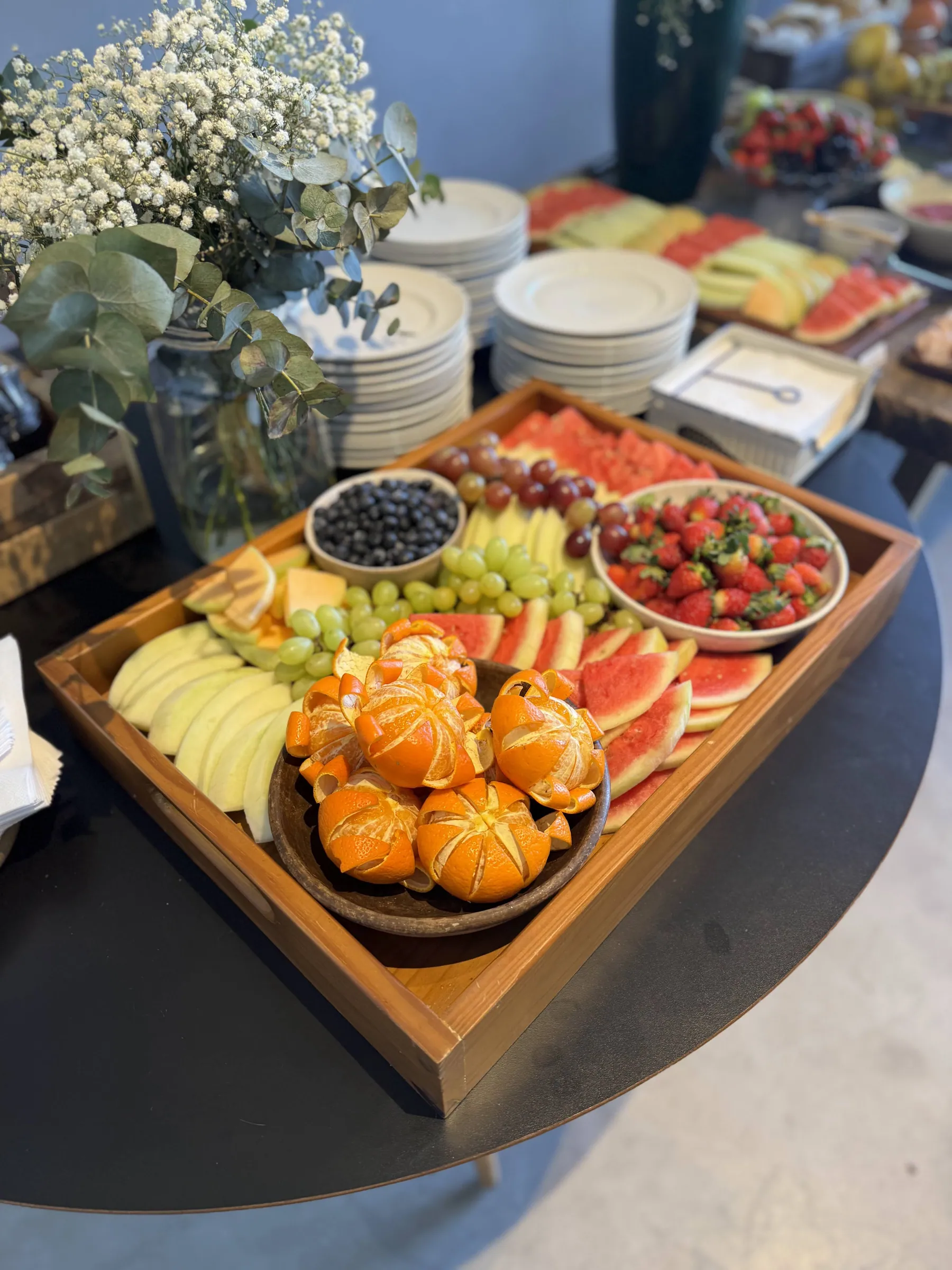 Bandeja de madeira farta com frutas variadas — melancia, uva, morango, mirtilo e mexerica descascada e cortada em pétalas — sobre uma mesa de evento com flores brancas ao fundo
