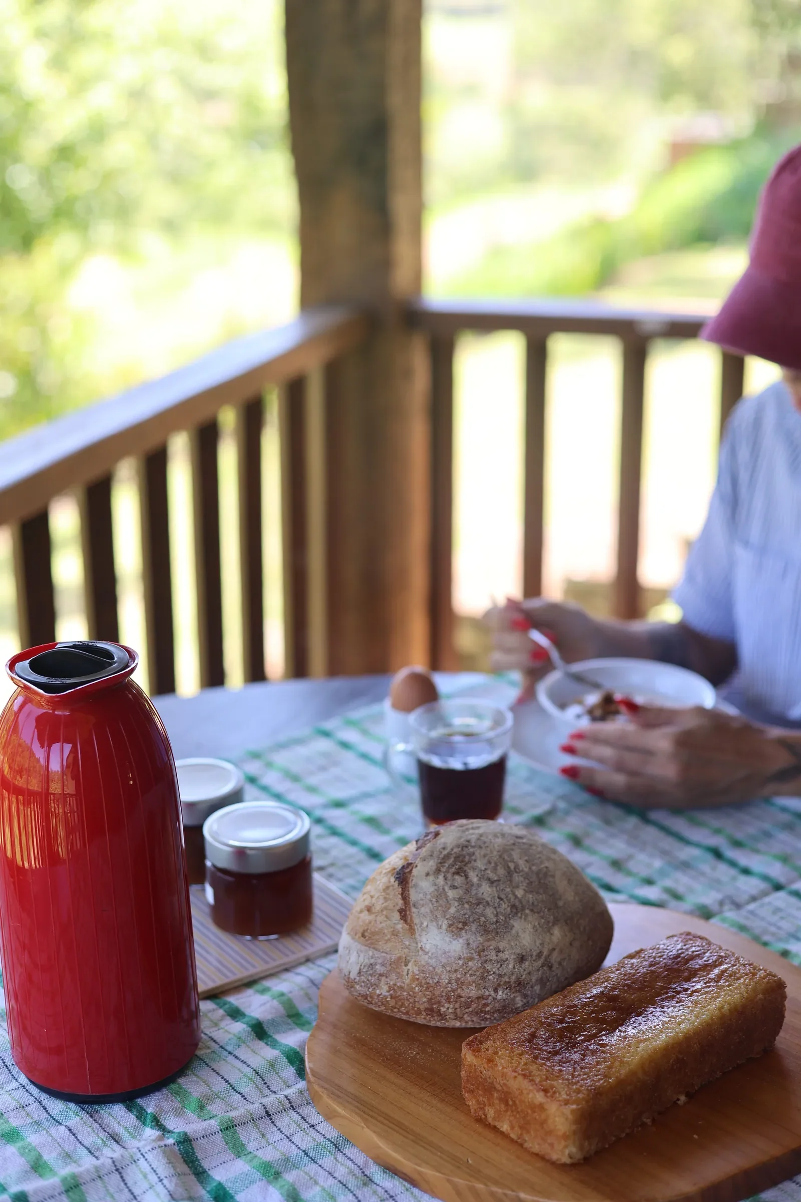 Mesa de café da manhã na varanda da Fazenda com garrafa térmica vermelha, pão, bolo e potes de geleia, vista para as montanhas ao fundo