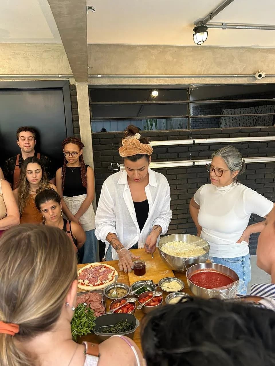 Chef explicando o preparo da pizza com as mãos sobre a massa, cercada por um grupo atento e tigelas de queijo e molho na bancada