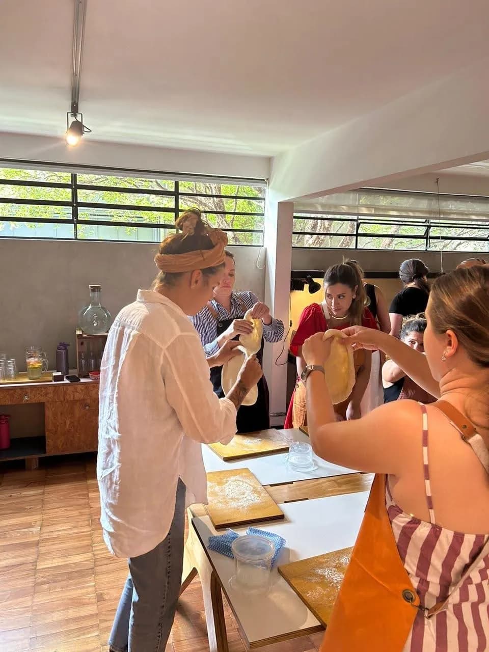 Grupo posicionando pizzas nos fornos ao ar livre com pás longas, num pátio com cactos e luz do dia