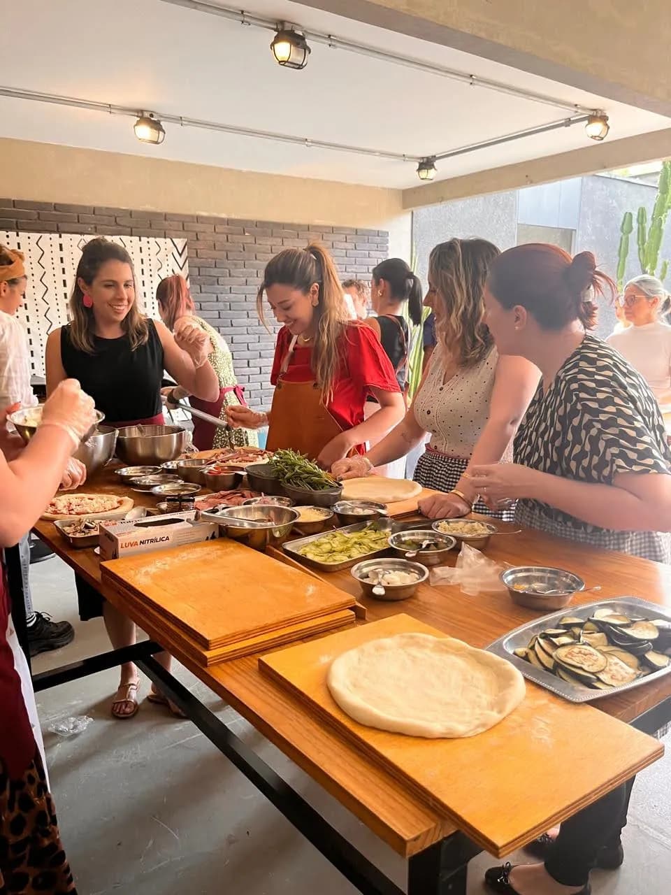 Grupo de participantes lado a lado numa mesa comprida, cada um sovando sua própria massa de pizza sobre tábuas de madeira, numa oficina mão na massa
