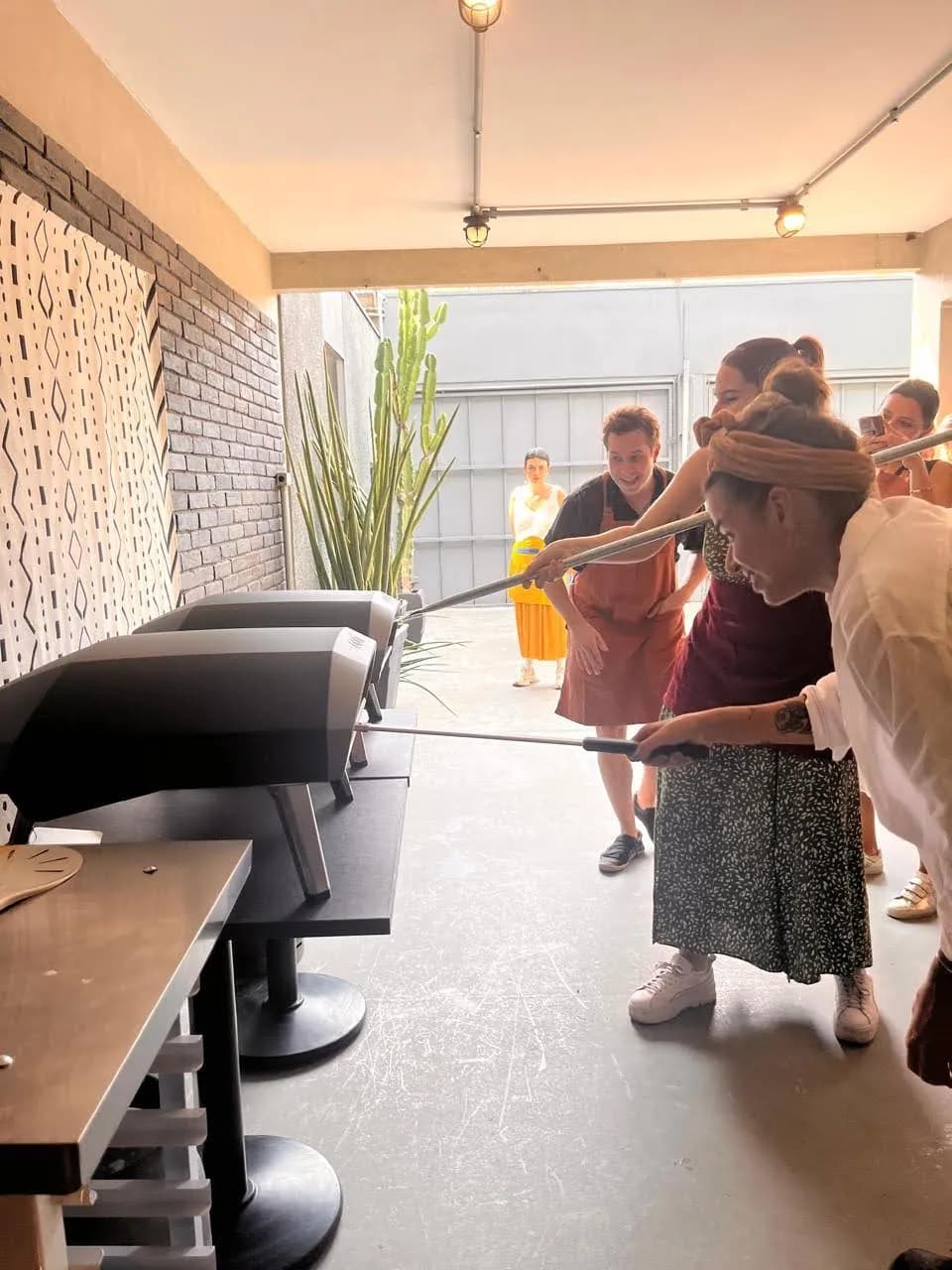 Duas pessoas esticando massa de pizza à mão, num ambiente iluminado por luz natural, com tábuas de madeira enfarinhadas ao fundo