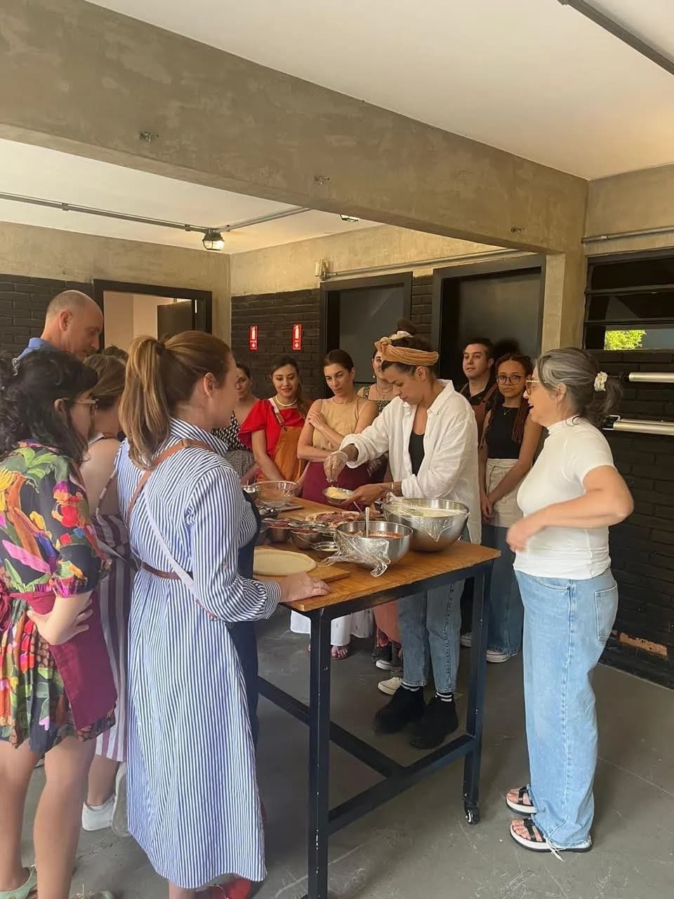 Grupo reunido ao redor de uma mesa de madeira com discos de massa já abertos e potes de recheios variados, atentos à explicação da chef