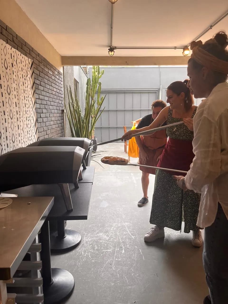 Participantes usando pás compridas para colocar pizzas nos fornos, em fila, observando o cozimento com curiosidade