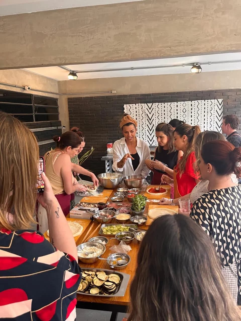 Chef de camisa branca montando uma pizza com salame e queijo diante de um grupo atento, com potes de molho de tomate e queijo ralado na mesa de madeira