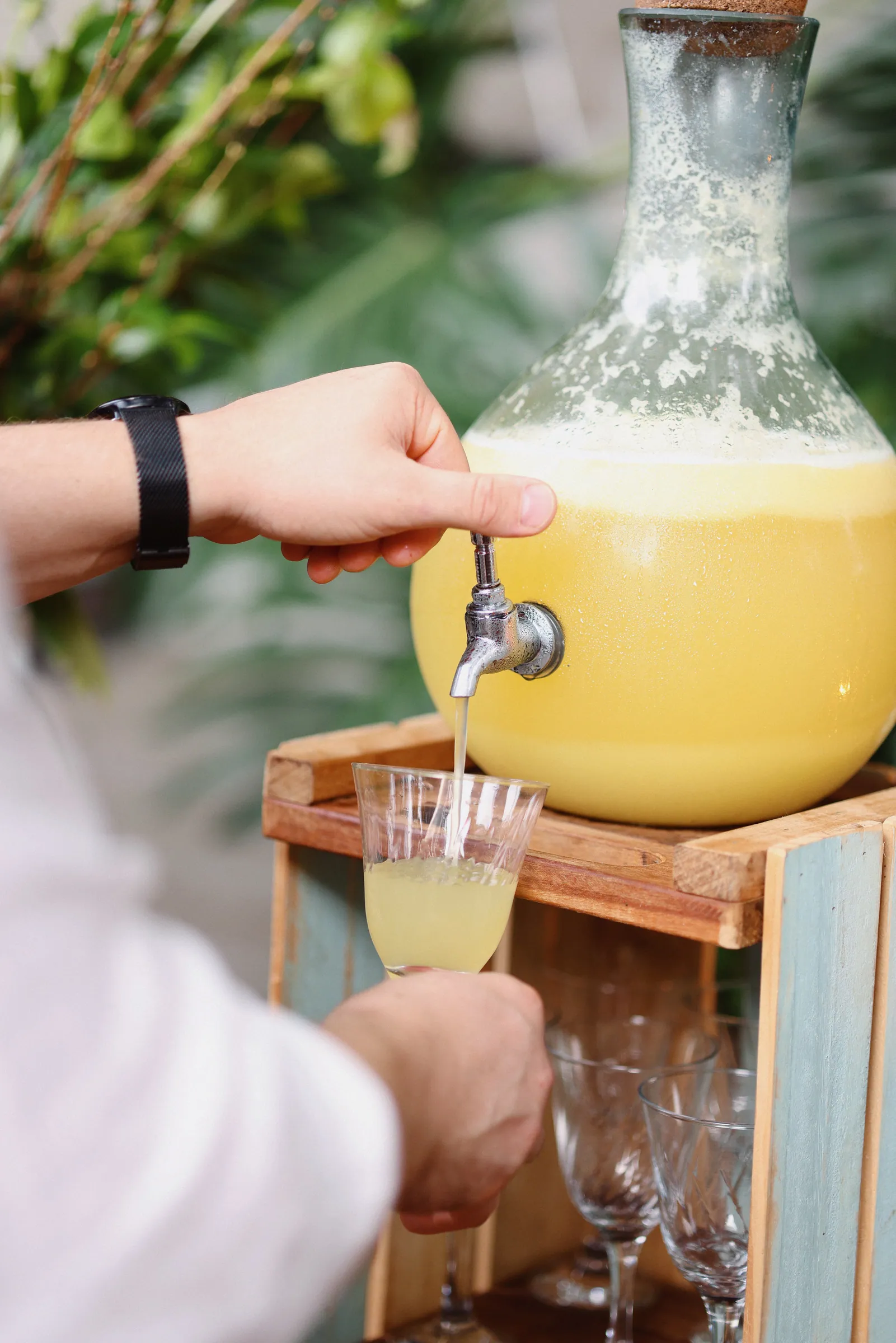 Mão servindo limonada de uma garrafa com torneira sobre suporte de madeira, copo se enchendo aos poucos