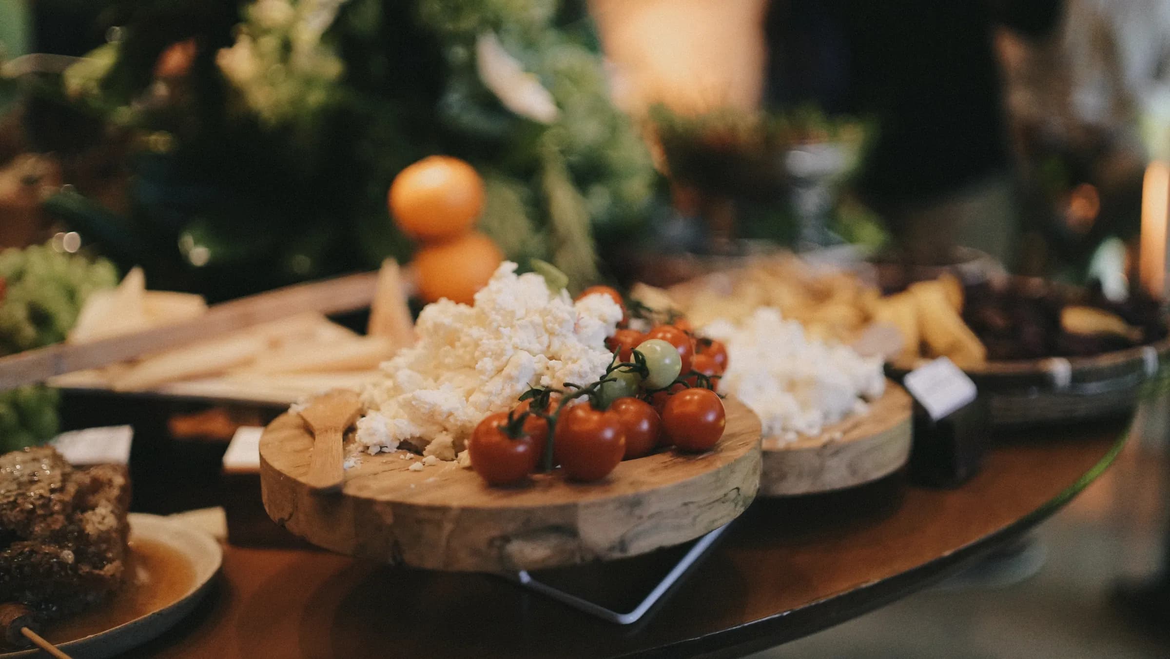 Queijo minas fresco e tomates-cereja sobre tábua de madeira, tangerinas e crackers ao fundo da mesa de frios