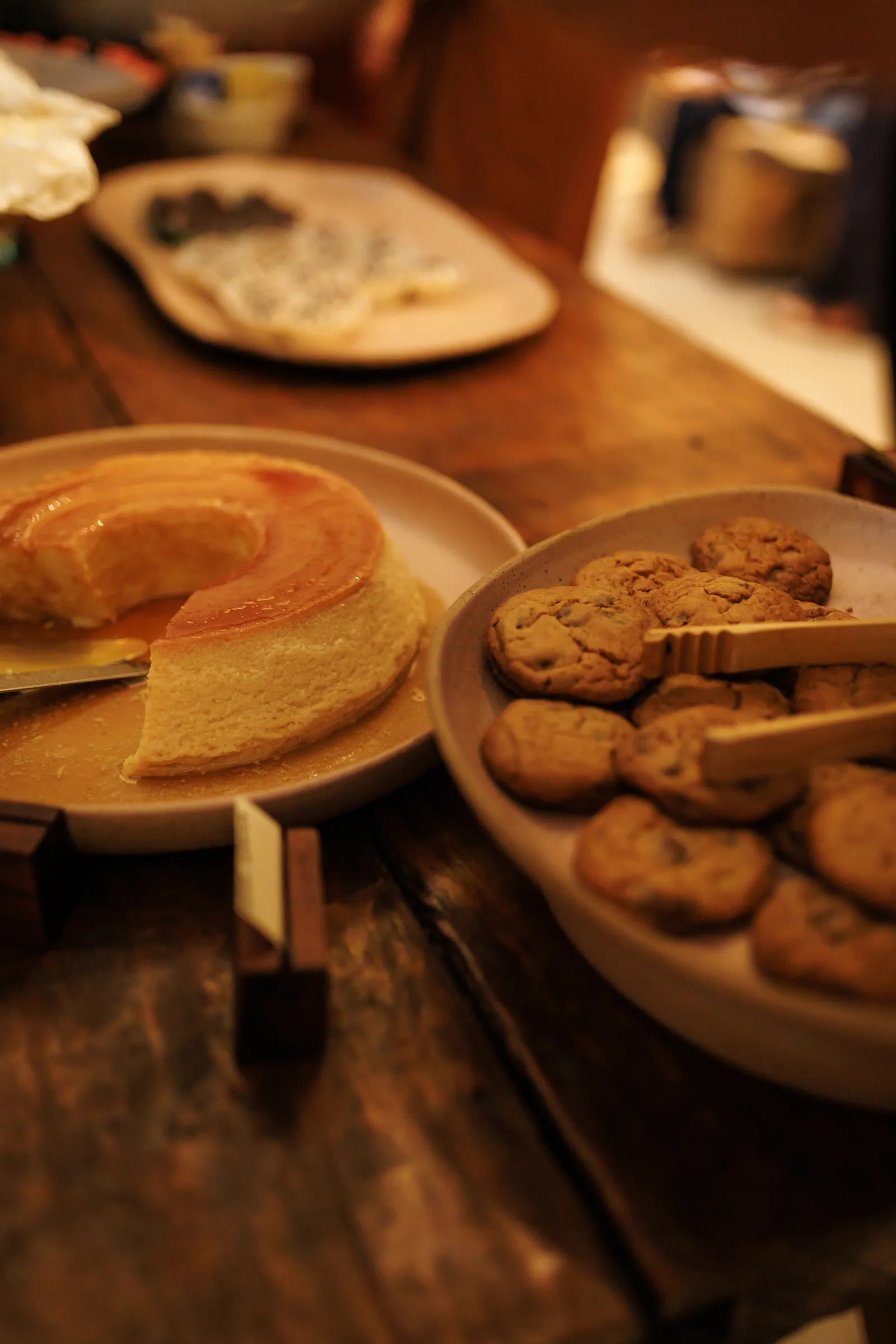 Pudim de leite fatiado ao lado de uma travessa de cookies, na mesa de doces com plaquinhas de identificação