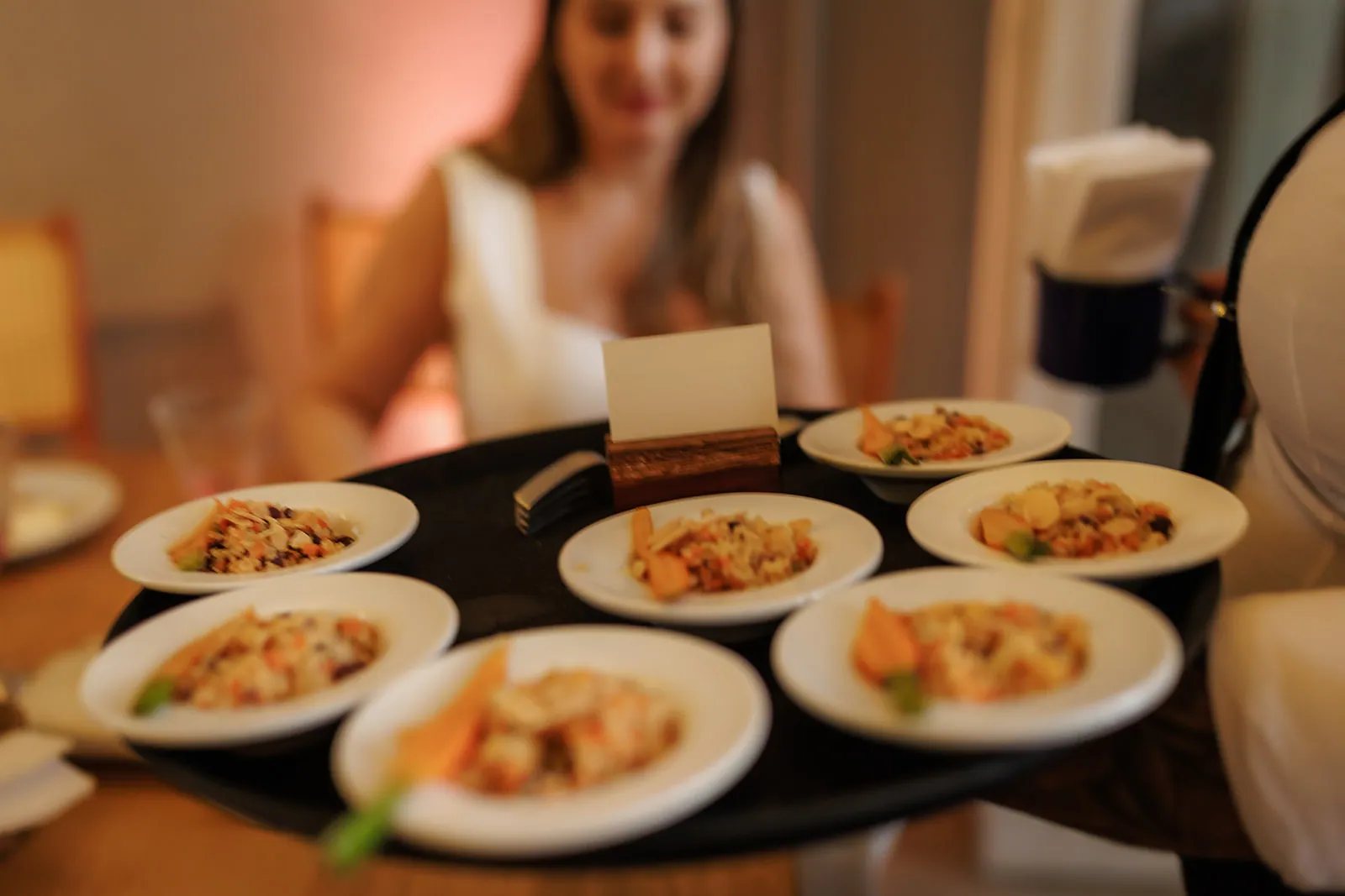 Garçom servindo pratinhos individuais de risoto com farofa numa bandeja redonda, convidada sorrindo à mesa
