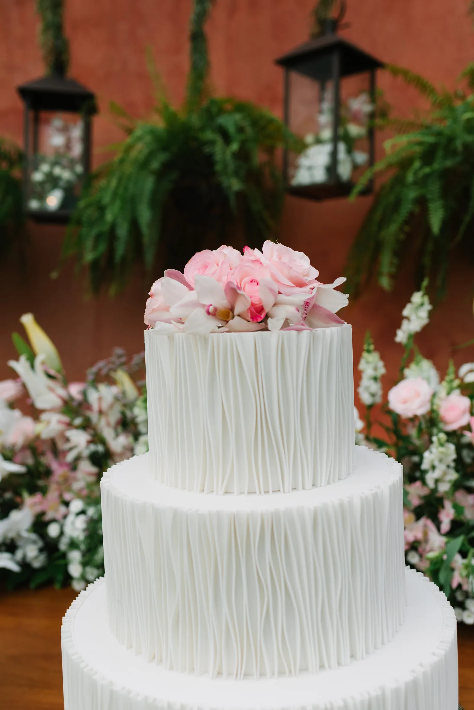Detalhe do bolo de casamento branco com textura ondulada e rosas claras no topo, luminárias penduradas ao fundo