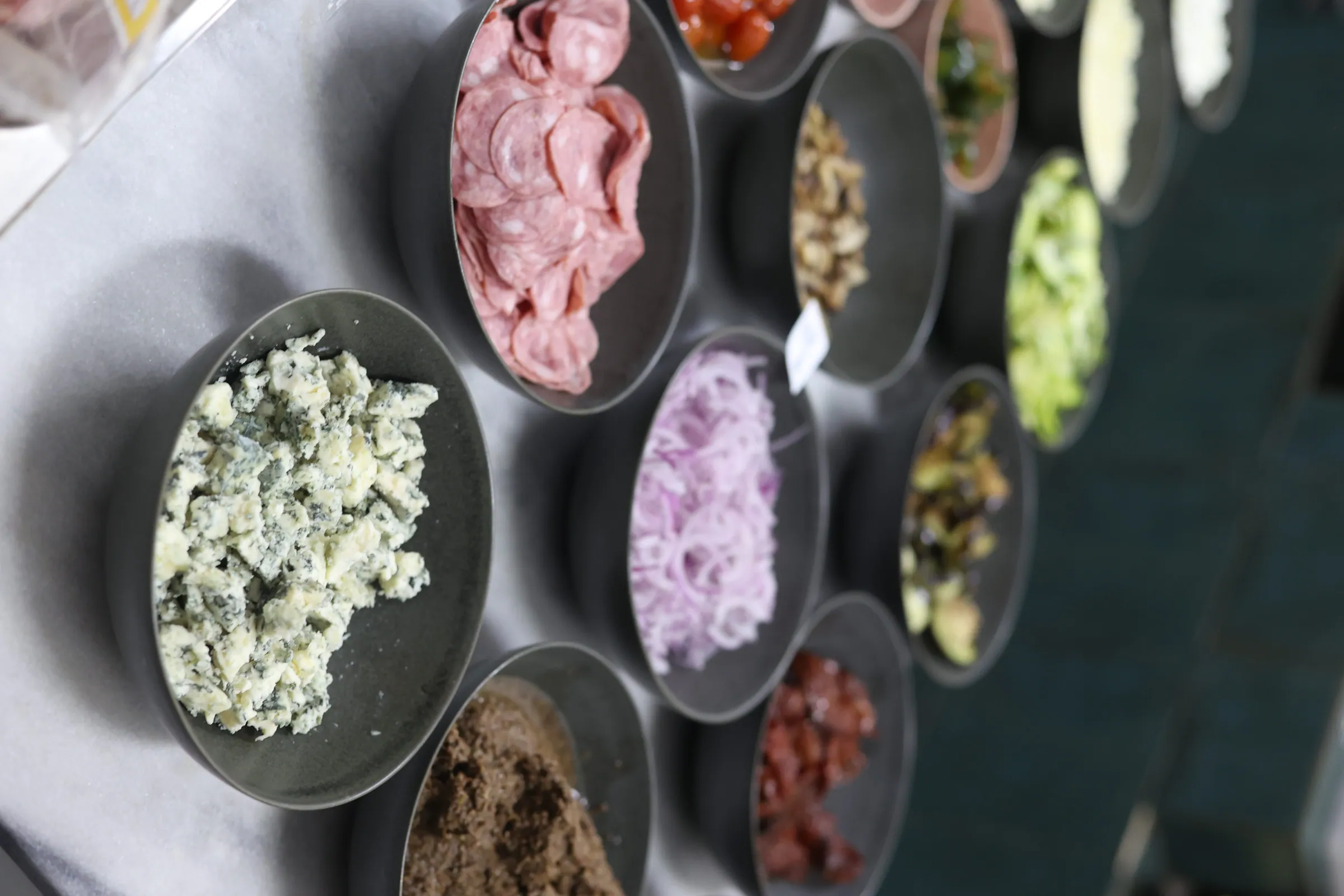 Estação de frios vista de cima, com potes de gorgonzola, salame fatiado, cebola roxa e outros acompanhamentos