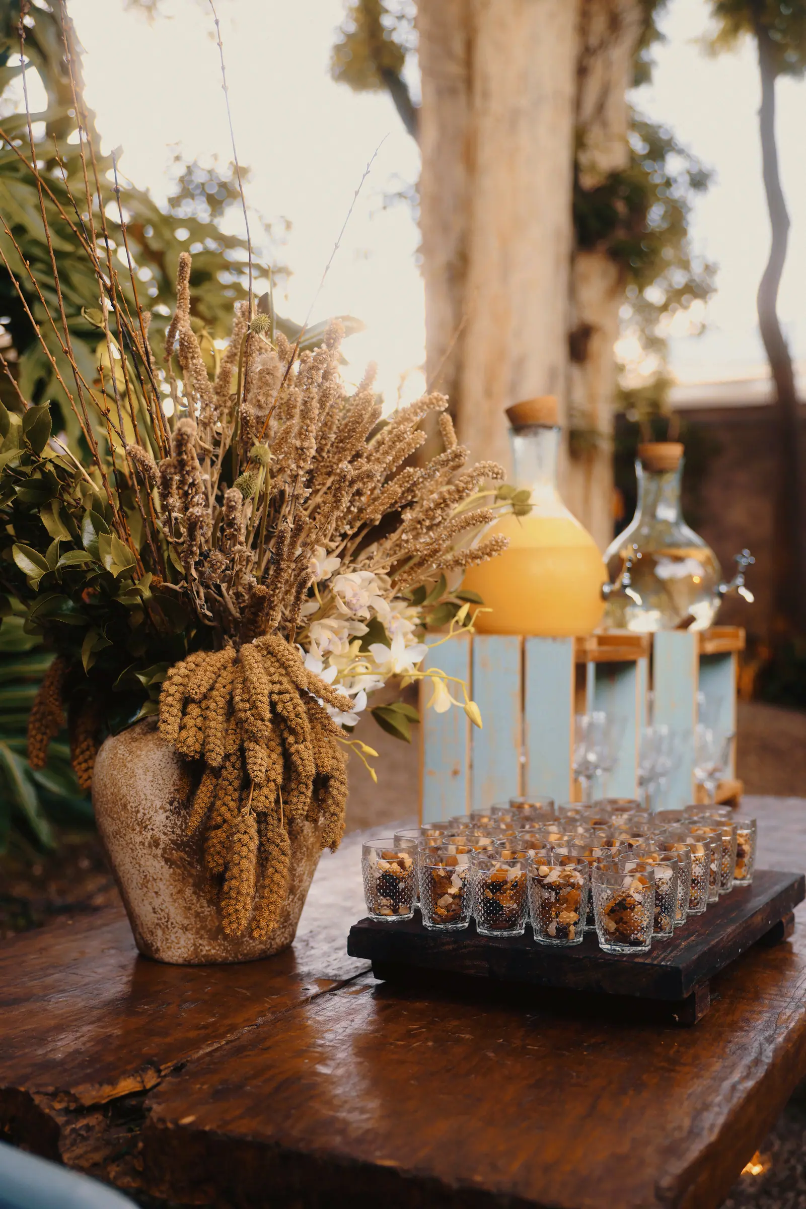 Drinks de boas-vindas em copinhos sobre mesa rústica, com garrafas de vidro e arranjo de flores secas ao lado