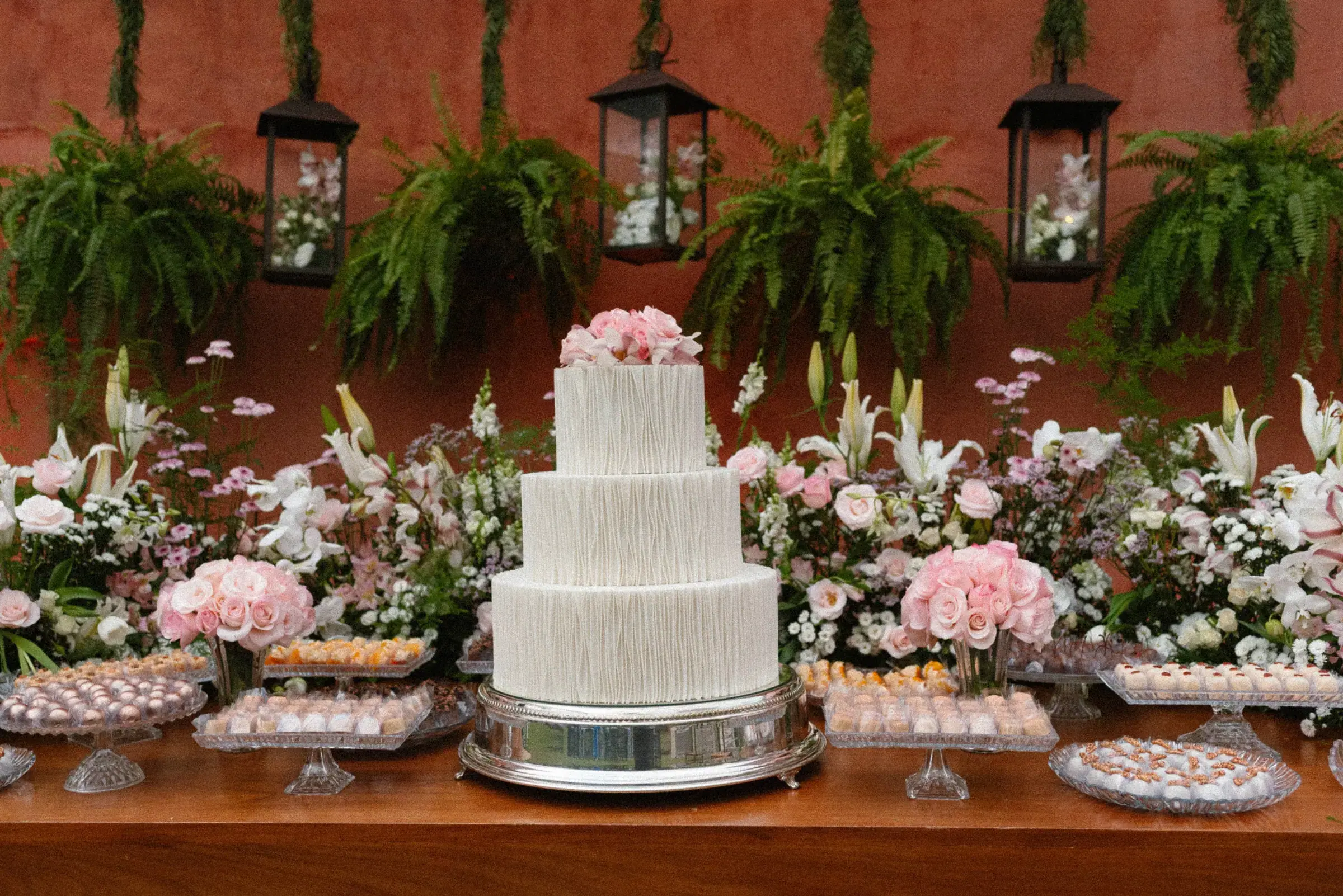 Bolo de casamento de três andares brancos com rosas claras no topo, cercado por travessas de docinhos e arranjos de flores diante de uma parede terracota