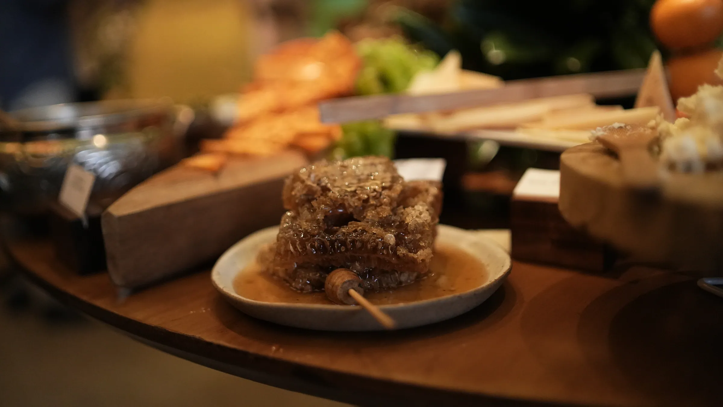 Favo de mel com mel escorrendo sobre um prato de cerâmica, cercado por tábuas de queijo e pães na mesa do buffet