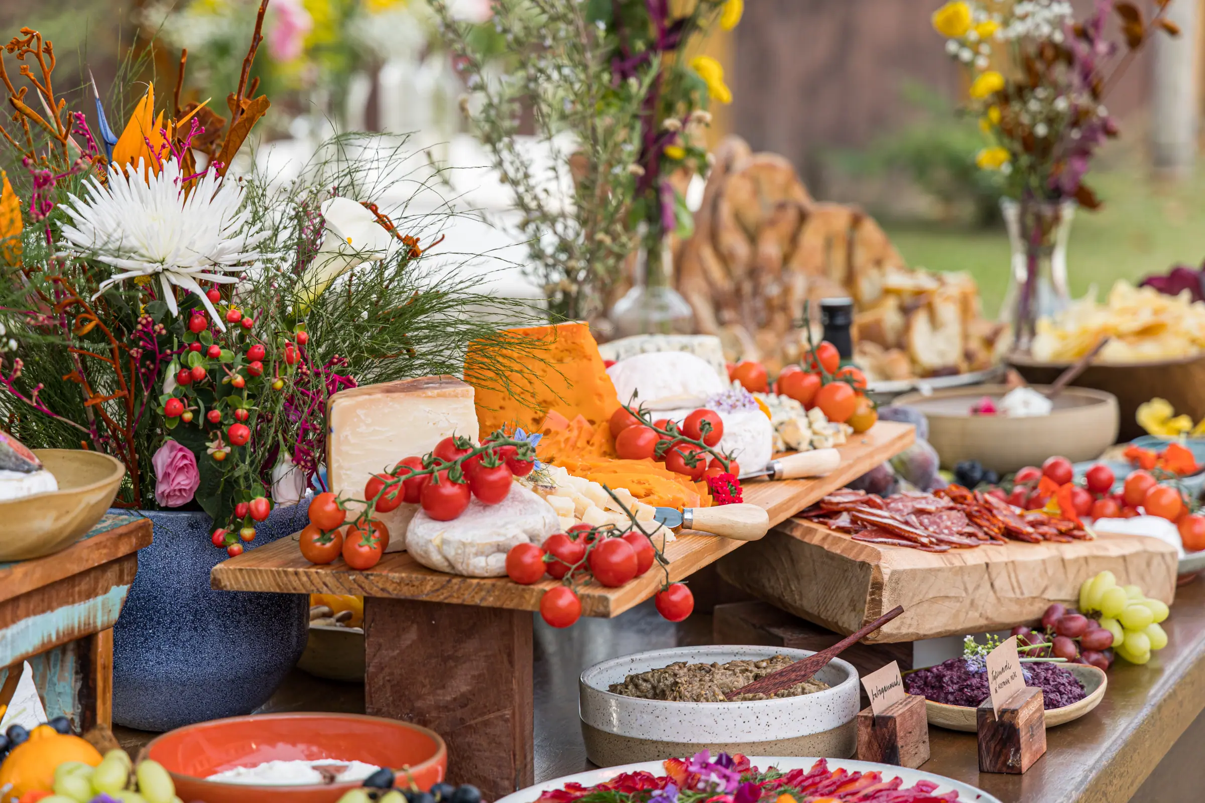 Mesa de antepastos em tábuas de madeira com tomates, queijos, embutidos e arranjos de flor do campo
