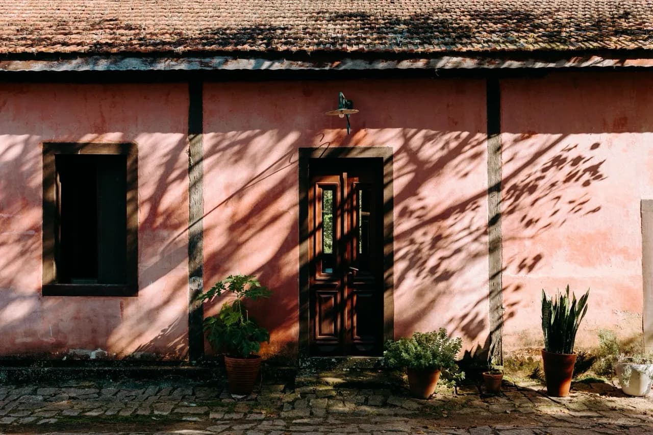 Parede rosa antiga da Fazenda Cósmica, com porta de madeira e sombras das folhas