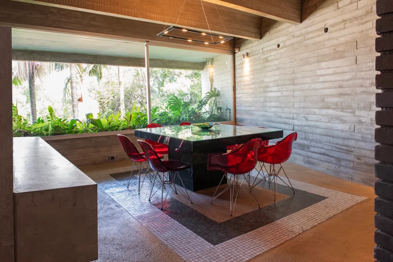 Nicho de jantar com mesa de pedra preta e cadeiras vermelhas transparentes, luminária pendente e vista para o jardim