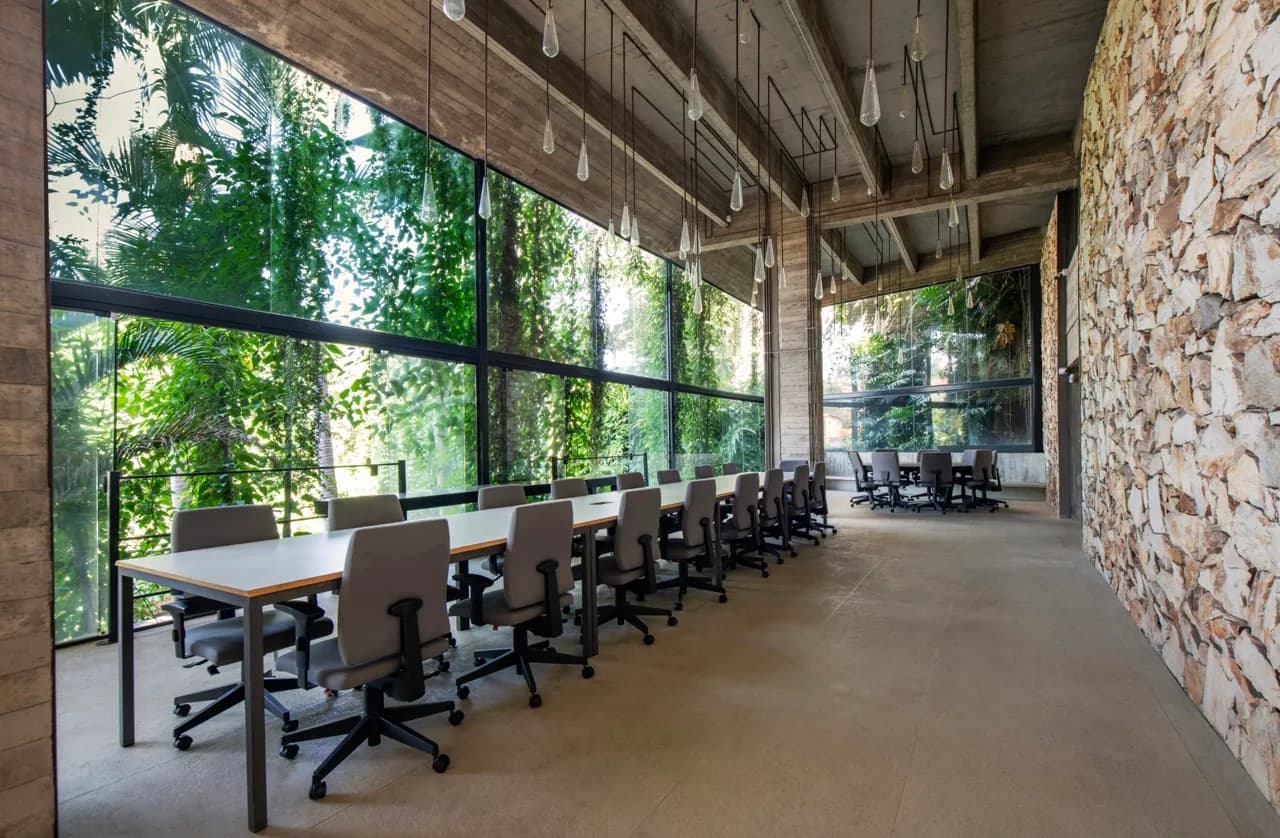 Sala de trabalho com mesas voltadas para a parede de vidro, mata fechando a vista e lâmpadas penduradas no teto