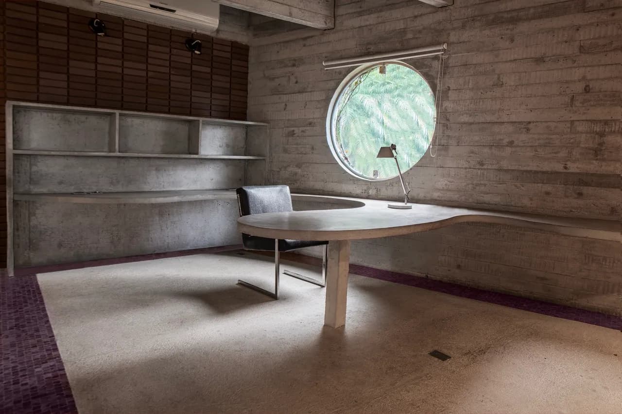 Mesa de trabalho orgânica sob janela redonda, estante de concreto ao fundo