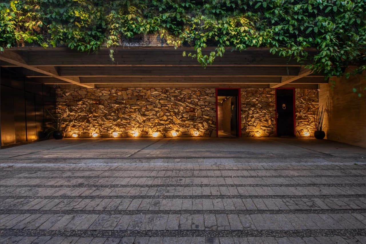 Entrada da Residência Cósmica à noite, parede de pedra iluminada por baixo entre trepadeiras