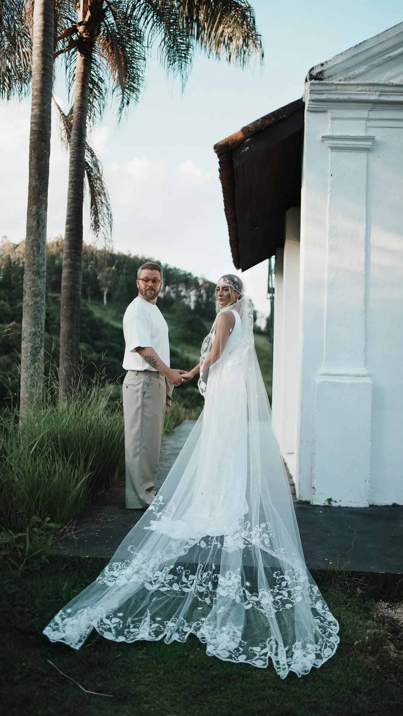 Casal de mãos dadas ao lado da capela da Fazenda Cósmica, com vestido longo e véu ao vento, palmeiras e morro atrás