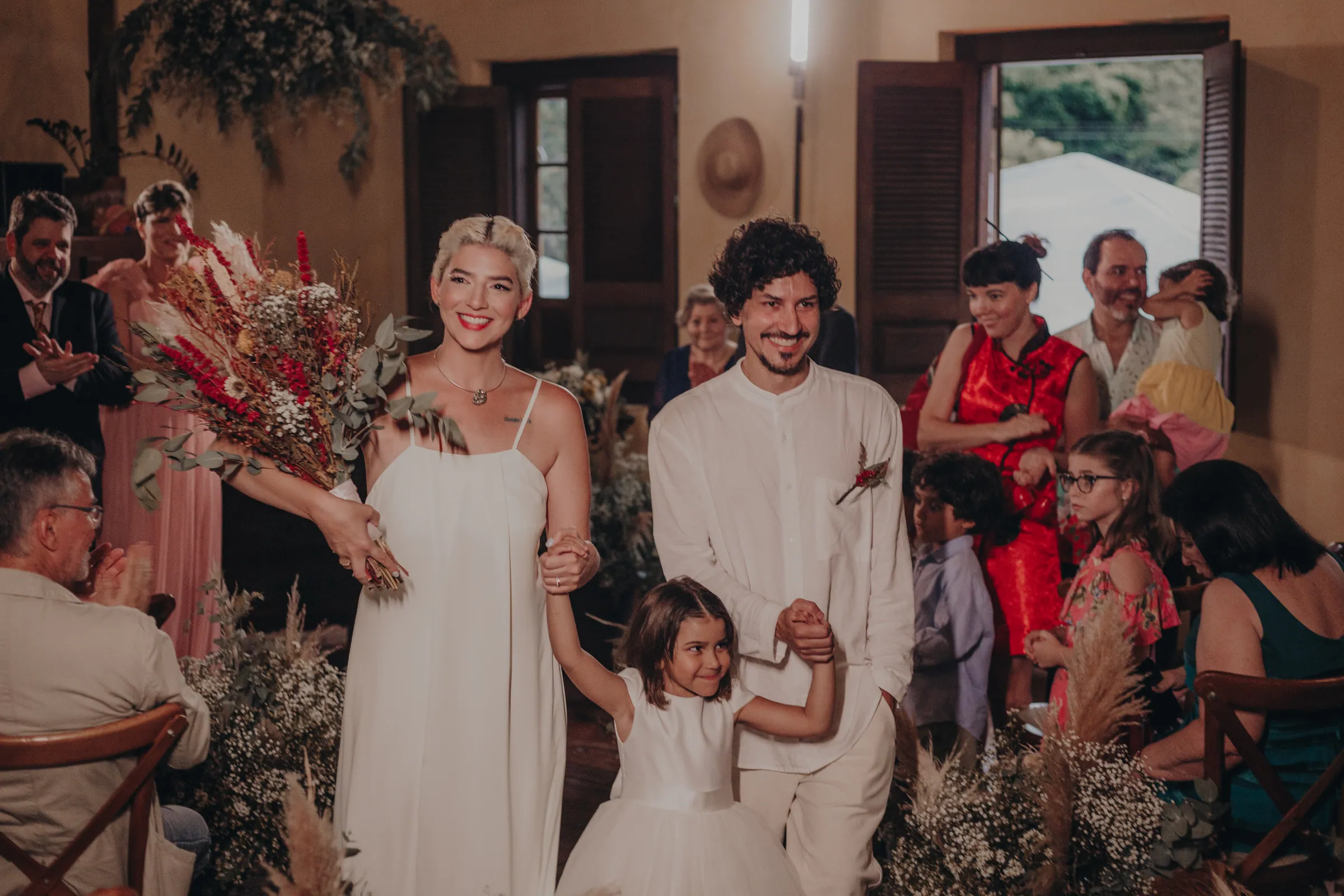 Casal recém-casado entrando no casarão de mãos dadas com a filha, convidados aplaudindo ao redor, decoração de flores secas vermelhas